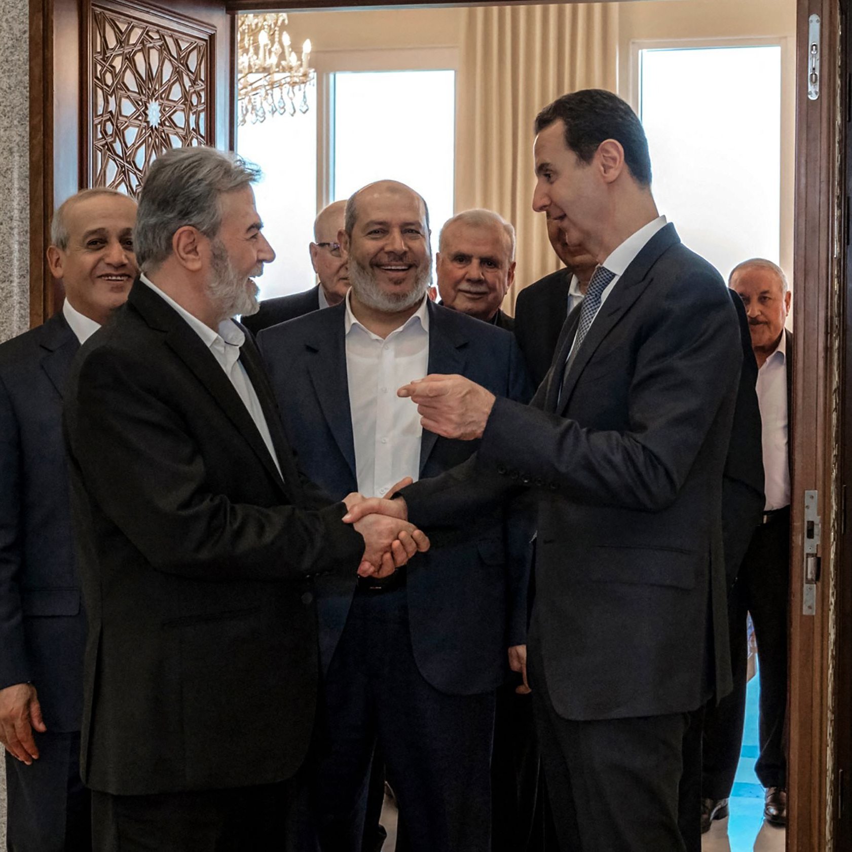 تظهر الصورة مجموعة من الرجال في قاعة استقبال. يتوسط الصورة شخصان يتصافحان، حيث يبدو أن أحدهما في وضعية حديث مع الآخر. الخلفية تحتوي على أبواب وزخرفة رائعة، وهناك أشخاص آخرون يقفون خلفهم، يظهر عليهم تفاعل إيجابي ورغبة في التواصل. الأجواء تبدو رسمية، مما يشير إلى لقاء مهم أو مناسبة خاصة.