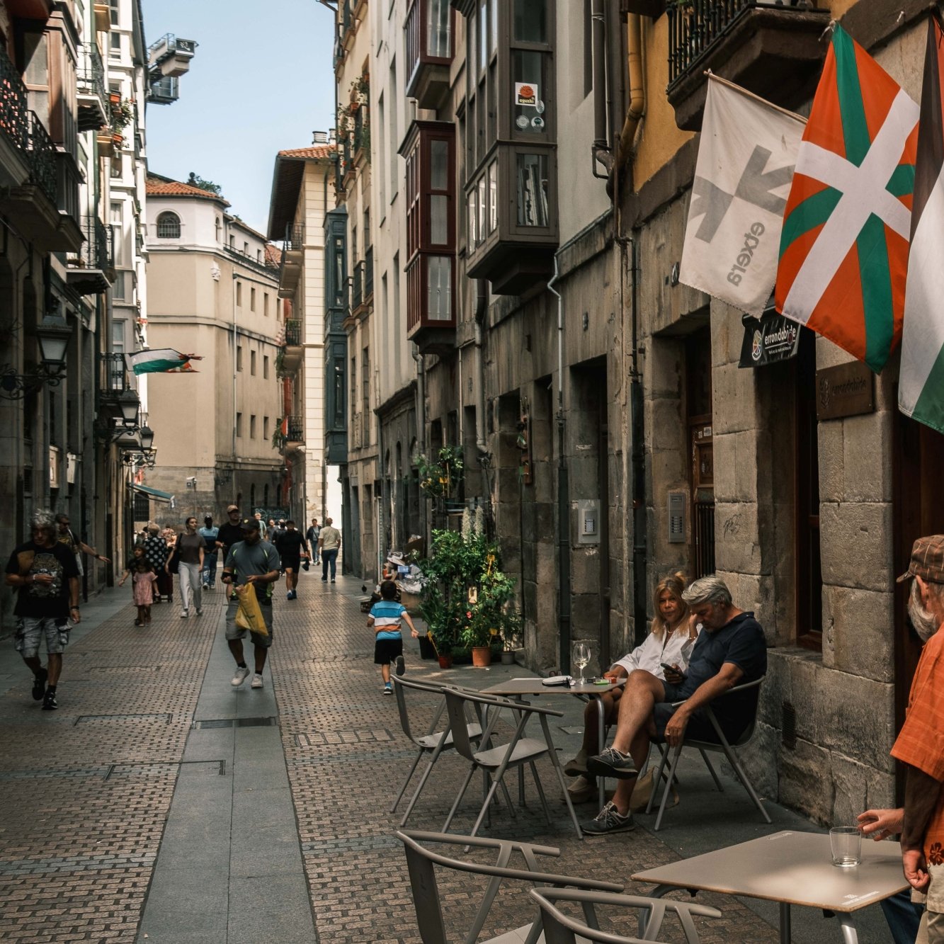 Calle estrecha con gente, mesas al aire libre y banderas ondeando en un ambiente urbano.