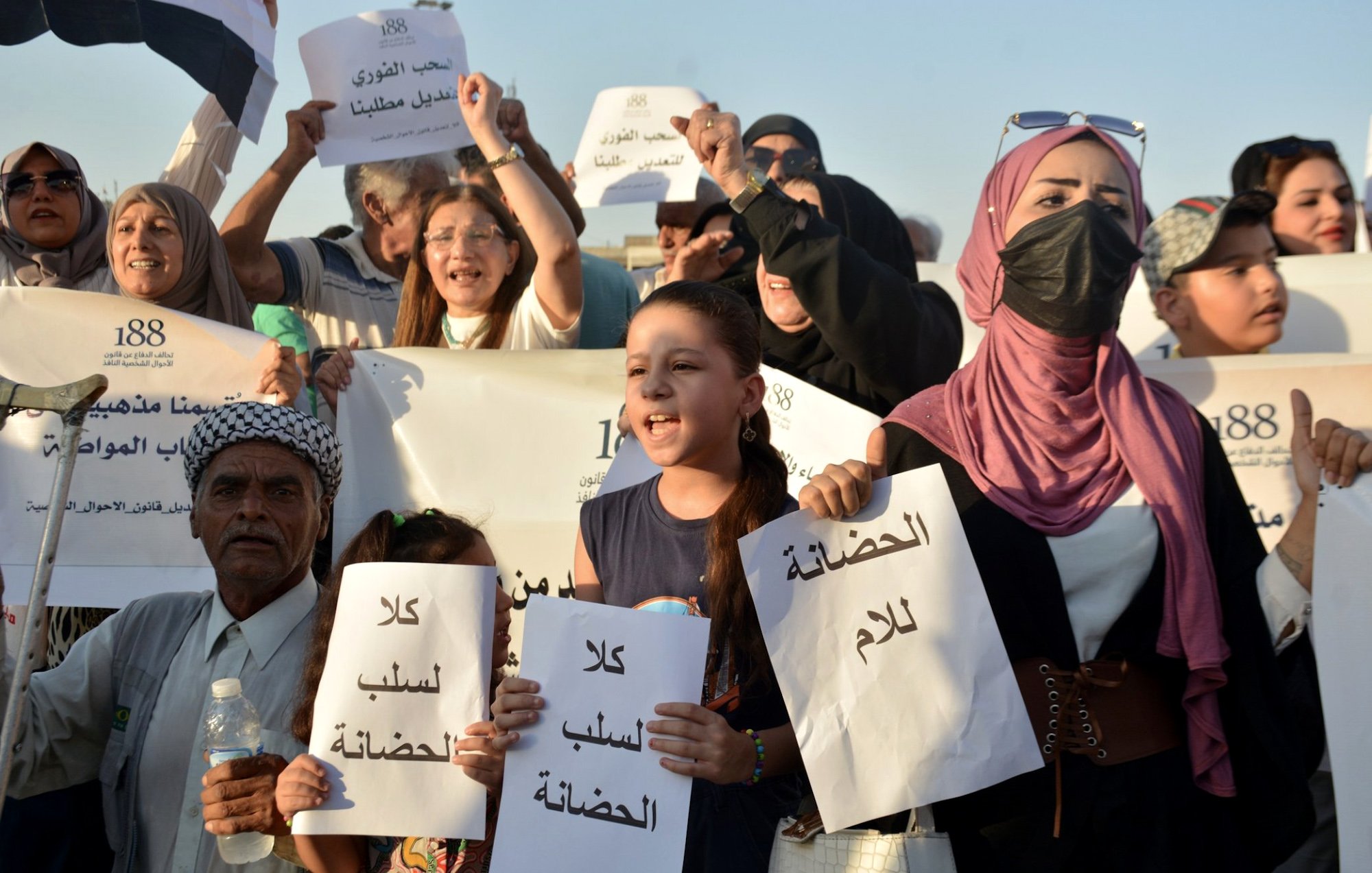 L'image montre un groupe de personnes lors d'une manifestation. Des manifestants, y compris des enfants, tiennent des pancartes avec des inscriptions écrites en arabe. Ils expriment des revendications, visiblement engagés dans une lutte pour une cause qui leur tient à cœur. Certaines personnes portent des masques, et l'ambiance semble déterminée et pleine d'énergie. Le paysage semble ensoleillé, suggérant un rassemblement en plein air.