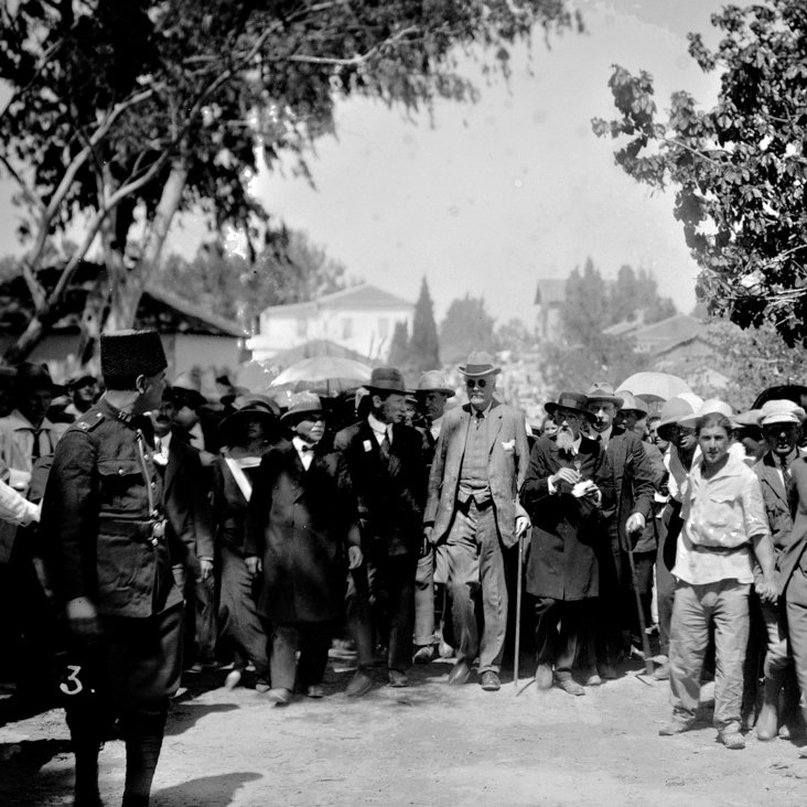 در این تصویر، گروهی از مردم در یک فضای باز دیده می‌شوند. بخشی از آنها چترهایی در دست دارند و ظاهراً در حال تماشای یک رویداد یا شخصیت مهم هستند. در وسط تصویر، مردی با عصا و لباس رسمی در حال حرکت است. اطراف او جمعیتی از مردان و زنان ایستاده‌اند که همگی لباس‌های رسمی و مرتب به تن دارند. در سمت چپ، فردی در لباس نظامی دیده می‌شود که به نظر می‌رسد به عنوان نگهبان یا محافظ عمل می‌کند. فضای عمومی و هوای آفتابی نشان‌دهنده یک روز شلوغ و پرحادثه است.