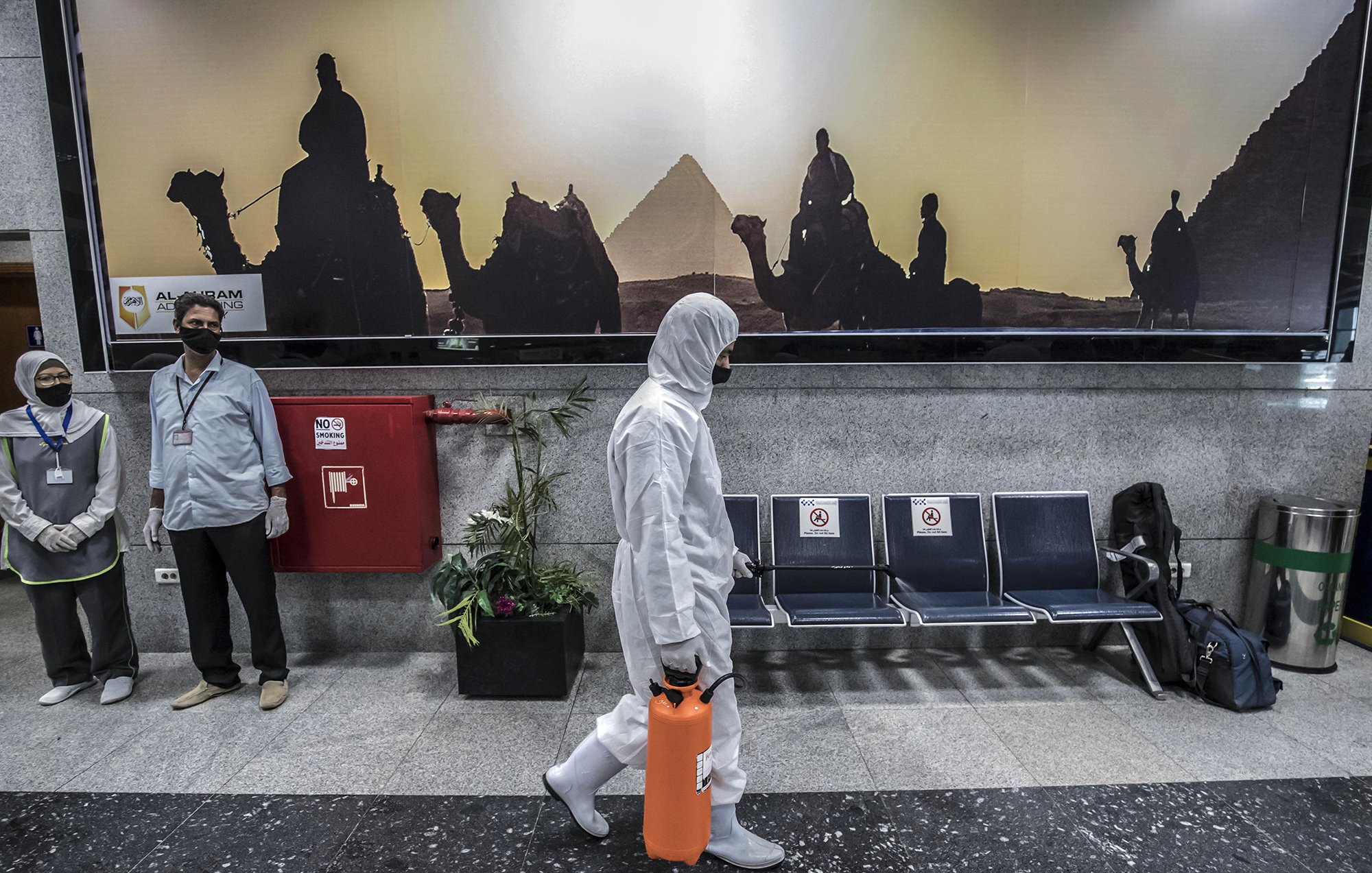 The image depicts a scene in a public area, likely an airport or a similar facility. In the foreground, a person wearing a white protective suit and mask is walking while holding an orange container. In the background, a large advertisement features silhouettes of camels and a pyramid, suggesting a connection to Egypt. There are two individuals in the background, also wearing masks, standing near a fire extinguisher. The seating area appears empty, adding to a sense of quietness in the environment.