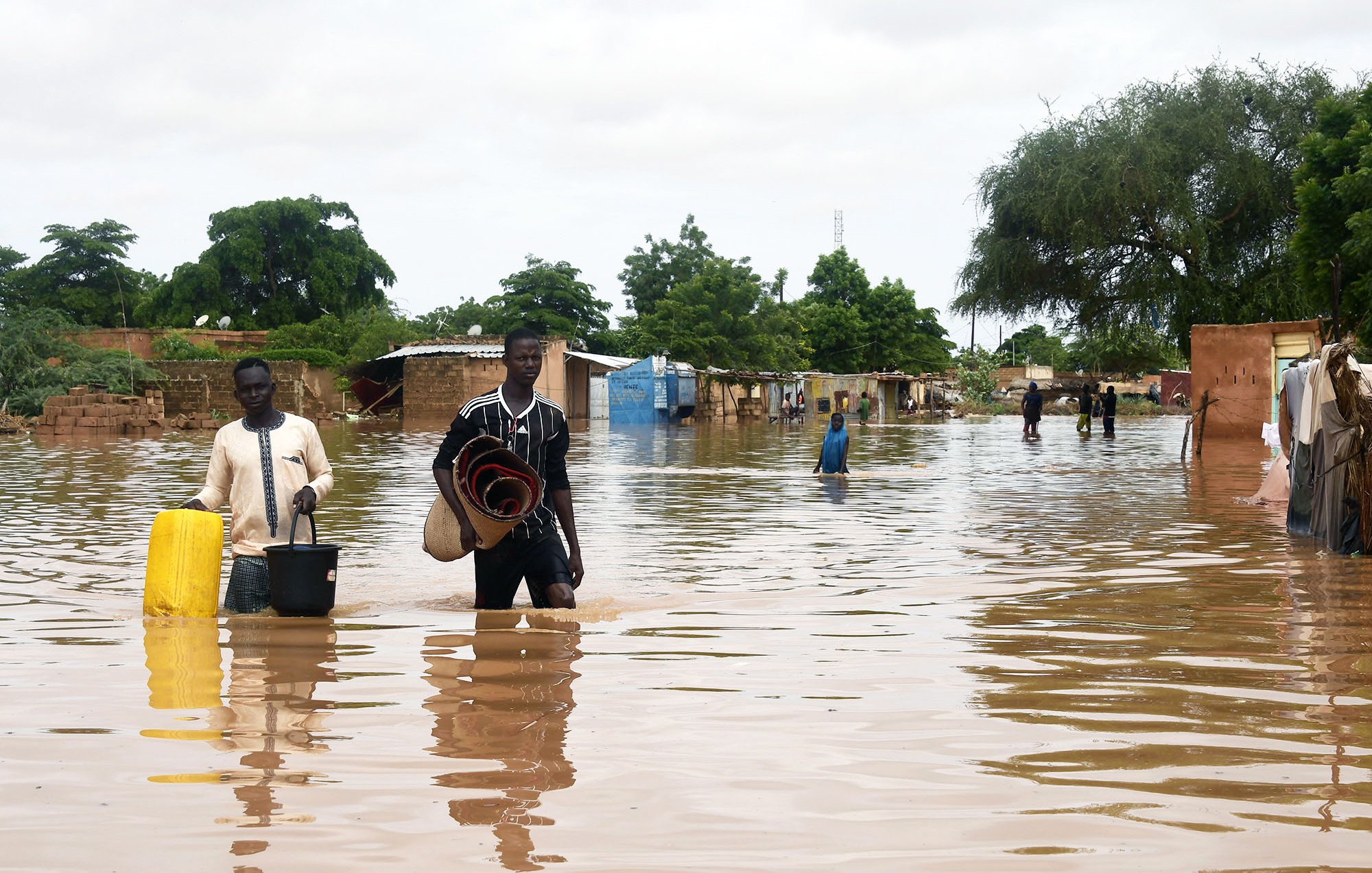 L'image montre une scène de inondation où deux hommes marchent dans une zone inondée. L'eau monte jusqu'à leurs jambes et environ une partie des maisons environnantes est submergée. Les hommes portent des objets, l'un avec un seau et l'autre avec des paniers ou des matelas. En arrière-plan, on peut voir des arbres et des bâtiments partiellement immergés, représentant une situation difficile due aux inondations. Le ciel est nuageux, ce qui suggère des conditions météorologiques instables.