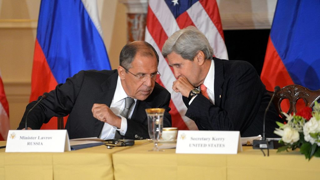L'image montre deux hommes assis à une table, engageant une conversation. L'un est identifié comme étant le ministre des Affaires étrangères de la Russie, Sergueï Lavrov, et l'autre comme John Kerry, l'ancien secrétaire d'État américain. Ils semblent discuter de manière confidentielle, entourés de décorations représentant les drapeaux de la Russie et des États-Unis. Sur la table, il y a des documents et une tasse. L'atmosphère semble sérieuse et diplomatique.