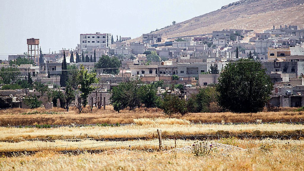 تظهر الصورة منطقة ريفية حيث يتواجد عدد من المنازل والمباني على تلة. في الخلفية، يمكن رؤية جبال منخفضة، بينما في المقدمة يوجد حقل جاف بألوان ذهبية. تبرز بعض الأشجار والنباتات هنا وهناك، مما يضفي لمسة من الخضرة على المشهد. كما يوجد خزان مياه في الجانب الأيمن من الصورة، مما يشير إلى سكان المنطقة. الجو يبدو مشمسًا مع أجواء صيفية.