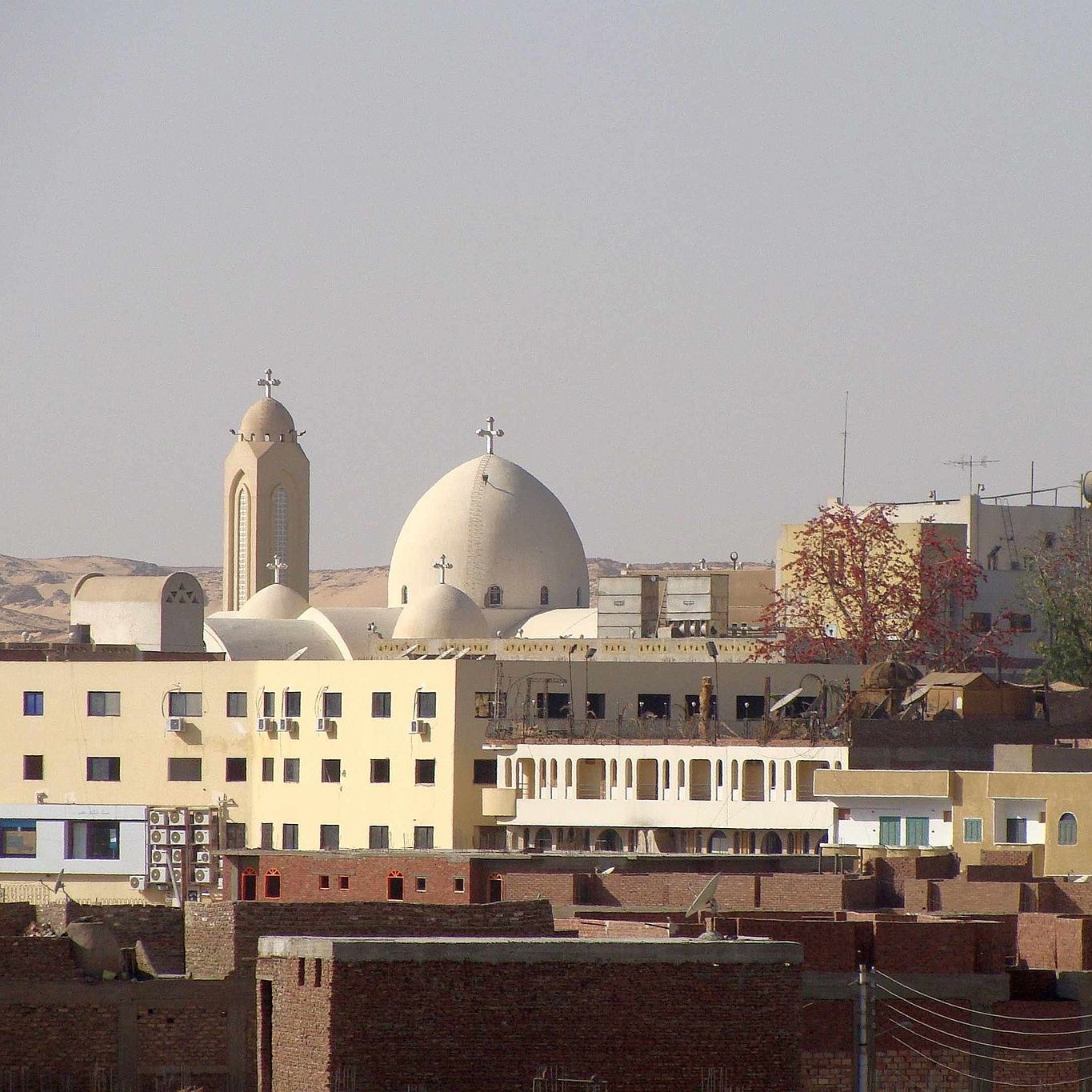 L'image montre un paysage urbain avec des bâtiments variés. On peut distinguer plusieurs églises avec des croix sur leurs dômes, caractéristiques de l'architecture religieuse. Des structures modernes cohabitent avec des bâtiments plus traditionnels, créant un contraste dans l'architecture. À l'arrière-plan, des collines ou des montagnes sont visibles, ajoutant une dimension naturelle au paysage. L'ensemble dégage une atmosphère de vie quotidienne dans une région marquée par son héritage culturel.