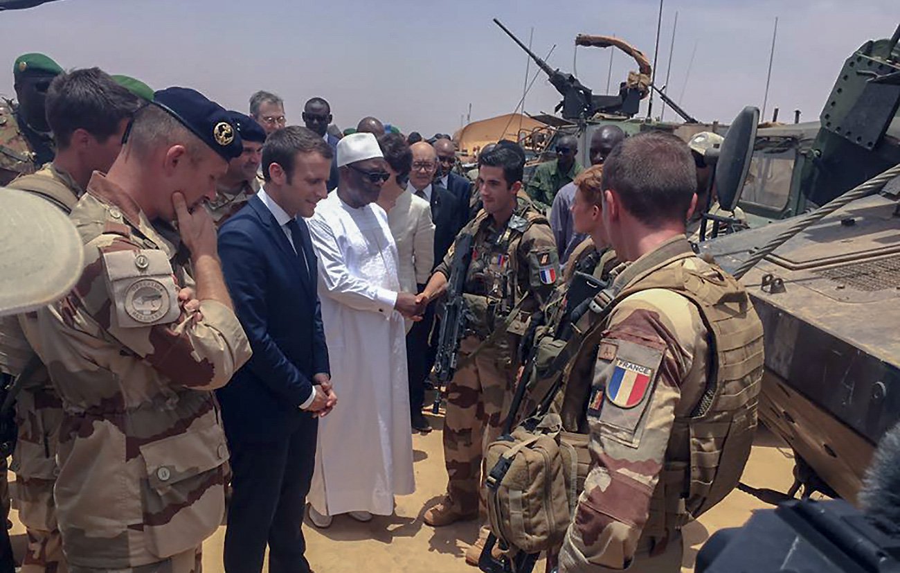 L'image montre un groupe de militaires et de responsables, probablement en situation de rencontre officielle dans un environnement désertique. Au centre, on peut voir un homme en costume, représentant vraisemblablement un leader, en train de discuter avec des soldats équipés de matériel militaire. Les militaires sont en uniforme et semblent engagés dans une mission, tandis que d'autres personnes obéissent à la dynamique de la réunion. L'atmosphère paraît sérieuse et solennelle.