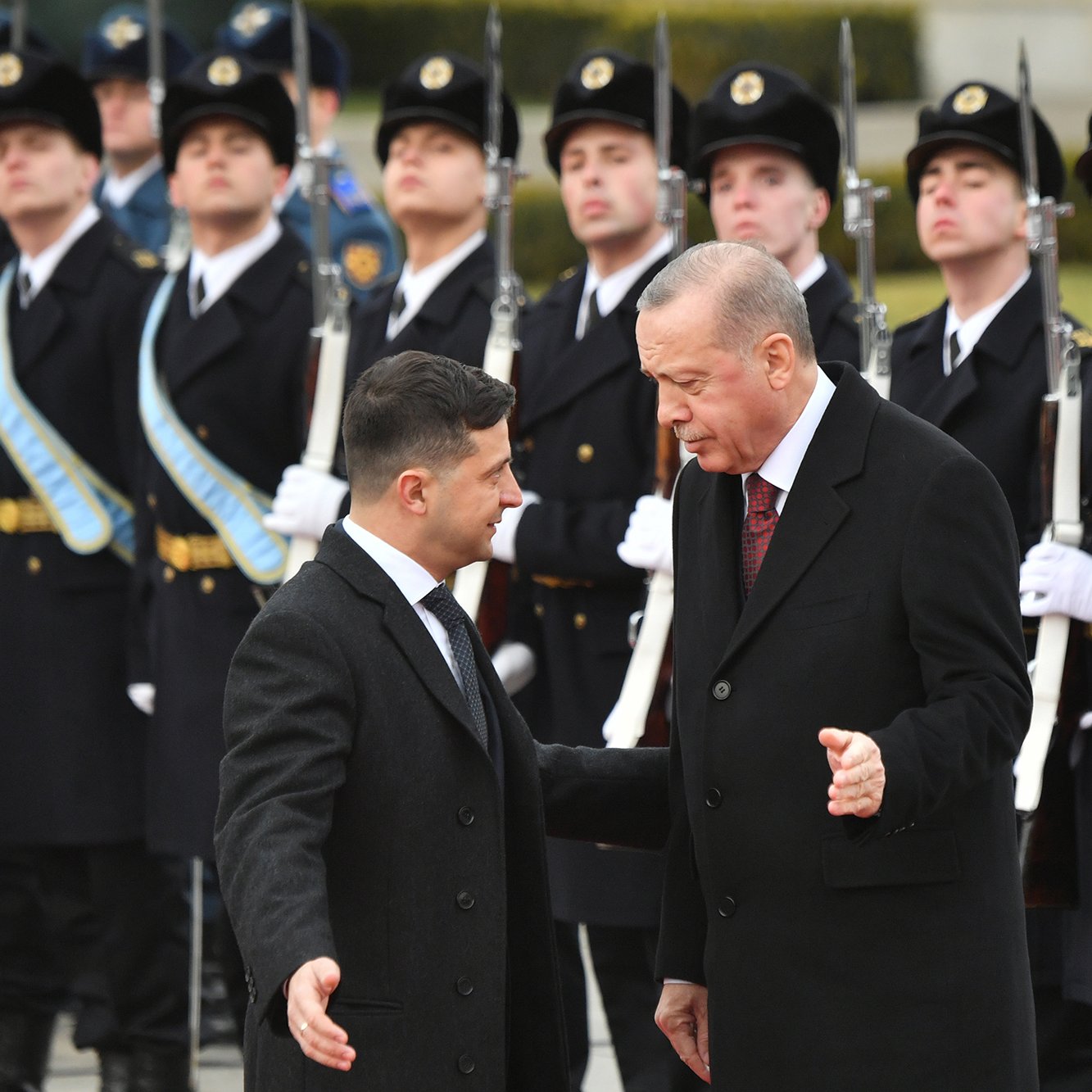 تظهر الصورة مجموعة من الجنود في الزي الرسمي يتواجدون في تشكيل عسكري أمام شخصيتين بارزتين. الشخصيات تبدو وكأنها تتحدثان معًا، حيث يبدو أحدهما في حالة نقاش مع الآخر. الجنود خلفهم يقفون بانضباط، بينما يتم عرض الأسلحة بشكل منظم. الأجواء تعكس طابعًا رسميًا وجديًا للحدث.