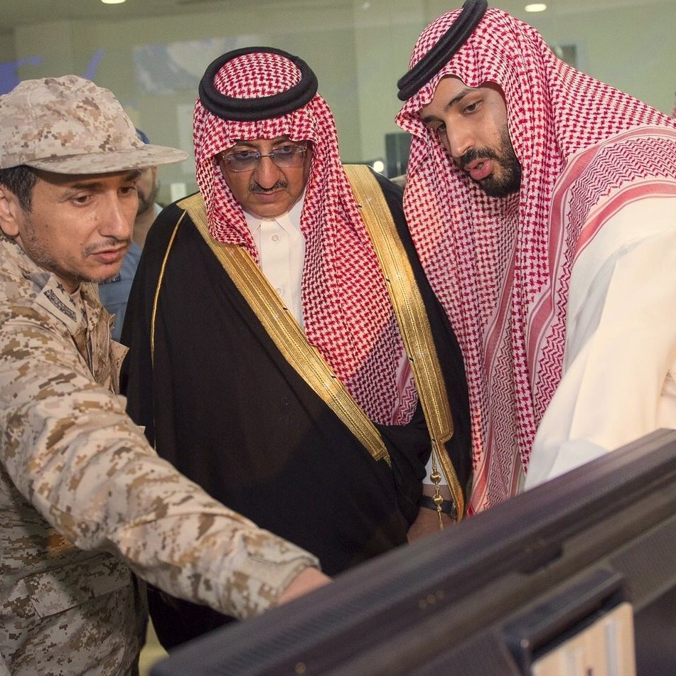 L'image montre un groupe de personnes, dont des hommes en tenue militaire et en vêtements traditionnels. L'un des hommes en uniforme semble expliquer quelque chose à ses collègues, qui sont attentifs à un écran d'ordinateur. L'environnement semble être un bureau ou une salle de réunion, et l'atmosphère suggère une discussion sérieuse ou une présentation. Les hommes semblent de haut rang, probablement impliqués dans des affaires militaires ou gouvernementales.