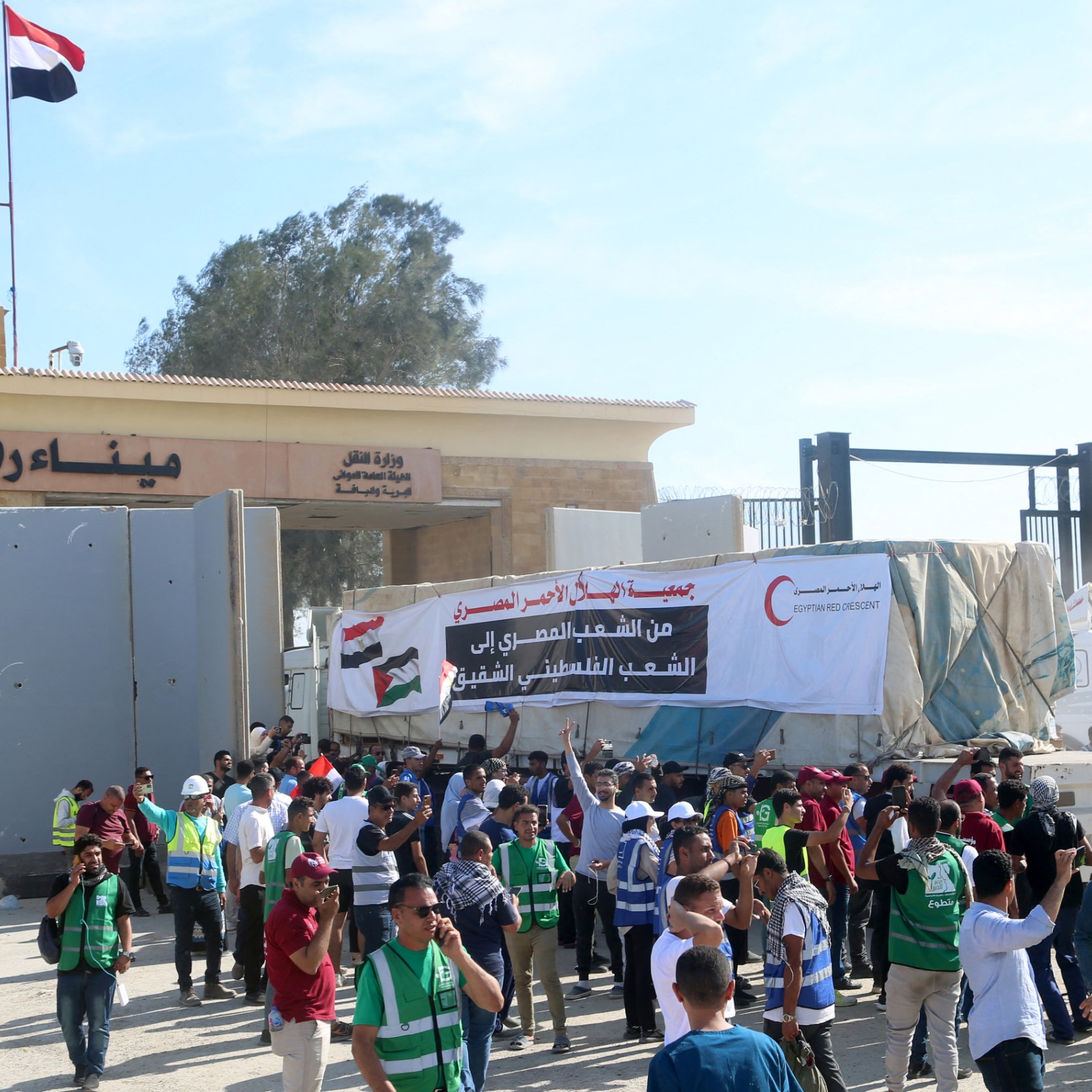 L'immagine mostra un gruppo di persone radunate davanti a un'entrata o a un confine, con bandiere nazionali visibili. Ci sono manifestanti che indossano gilet verdi e alcuni stanno tenendo striscioni. L'atmosfera sembra essere di protesta o di celebrazione, con diversi individui che interagiscono tra loro e partecipano all'evento. Sullo sfondo si vedono strutture architettoniche e una grande scritta in arabo.