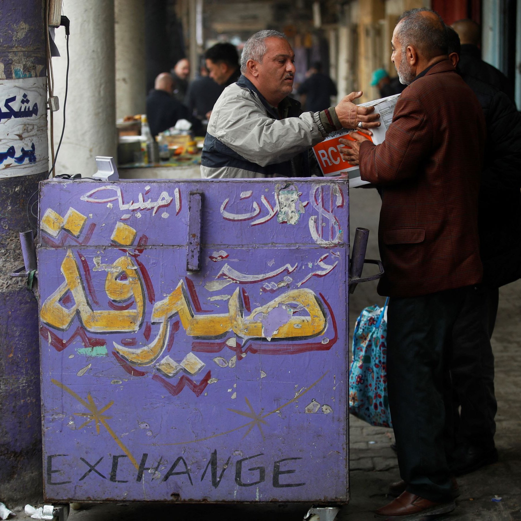 L'image montre un commerce dans une rue animée. On y voit un homme en train de discuter avec un autre, probablement en train d'échanger de l'argent ou des informations. En arrière-plan, des personnes sont assises à des tables, et on peut voir des étals remplis de marchandises. Le mur est décoré de graffitis et d'inscriptions en arabe, ainsi qu'une mention en anglais "EXCHANGE", ce qui indique probablement qu'il s'agit d'un bureau de change. L'ambiance semble vivante et typique d'un marché ou d'un bazar.