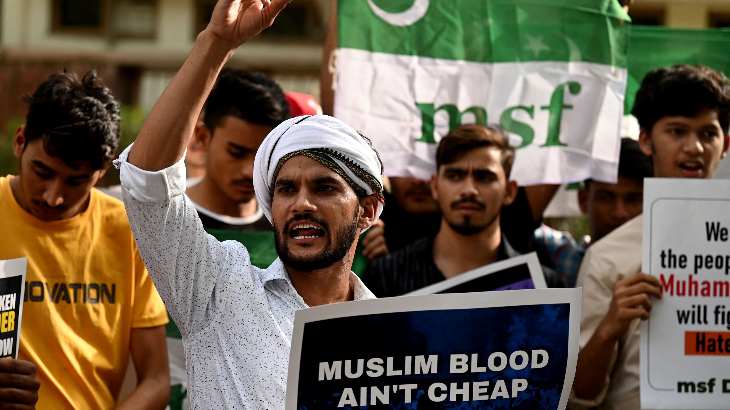 L'image montre un groupe de manifestants rassemblés, tenant des pancartes et des drapeaux. Au premier plan, un homme s'exprime avec passion, levant un doigt en l'air. Il porte une chemise claire et un turban. Les autres participants dans le fond semblent également engagés dans la manifestation, brandissant des affiches avec des messages. L'atmosphère semble intense et revendicative.