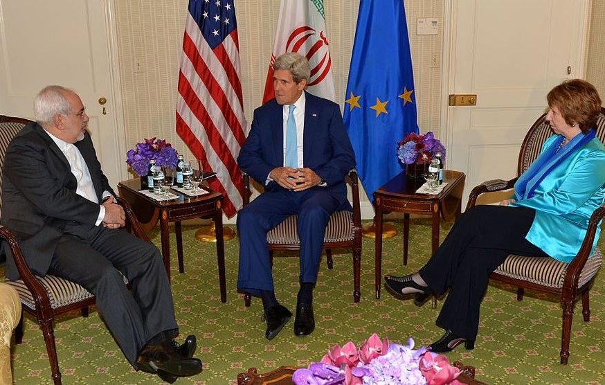 L'image montre une réunion en intérieur entre trois personnes assises dans un environnement élégant. À gauche, on voit un homme en costume noir, probablement un représentant de l'Iran. Au centre, un homme en costume clair, probablement un diplomate américain, semble discuter. À droite, une femme en blouse bleue, possiblement une diplomate européenne, écoute attentivement. Les participants sont entourés de tables avec des bouteilles d'eau et des fleurs en centre de table. L'atmosphère semble être celle de discussions diplomatiques sérieuses.