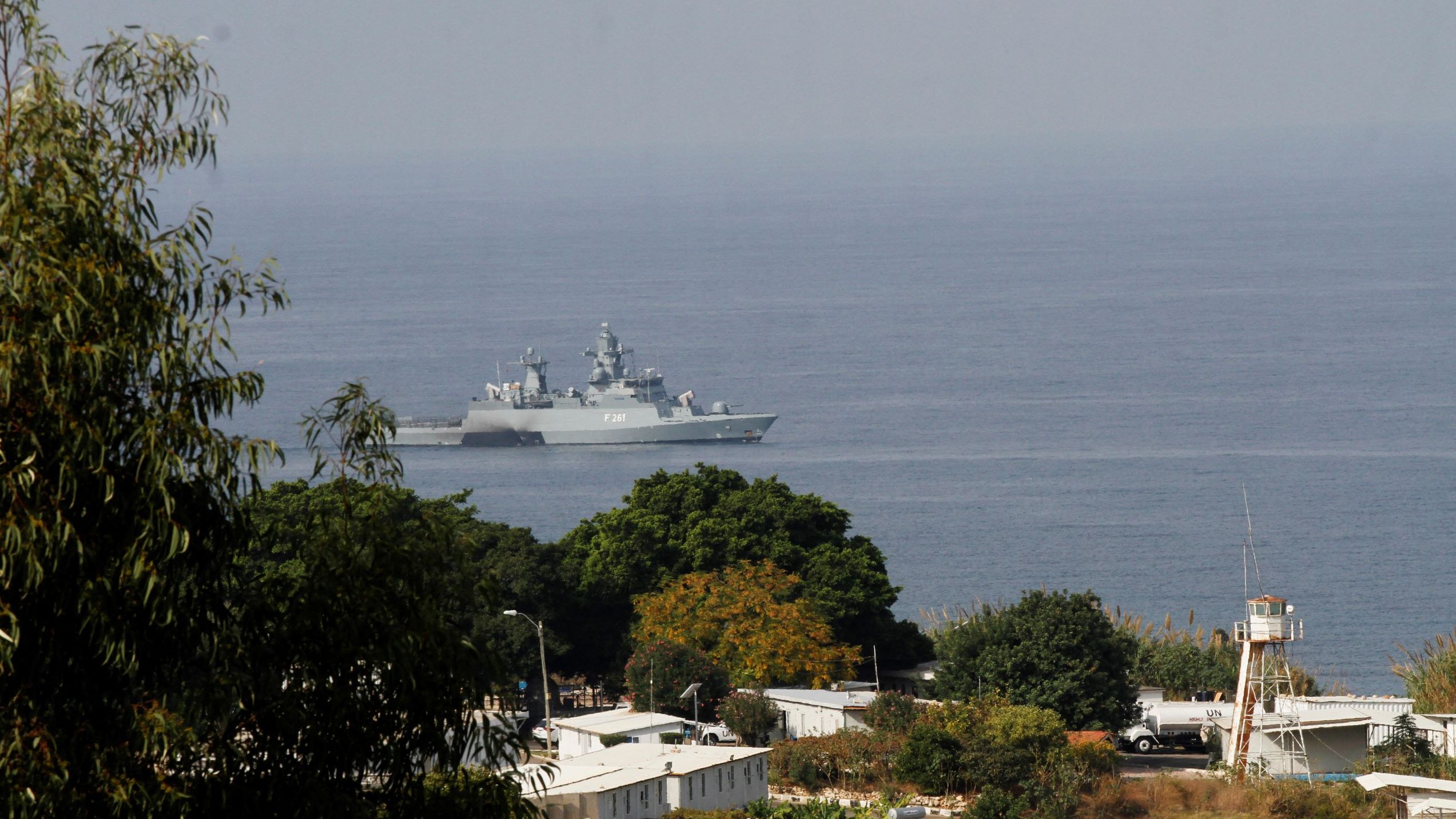 L'image montre un navire militaire se trouvant dans une étendue d'eau, probablement en mer. On aperçoit des paysages côtiers avec quelques arbres et des bâtiments en arrière-plan. Le navire a une structure moderne et est visible au loin, tandis que la végétation au premier plan donne une impression de profondeur à la scène. L'atmosphère semble claire, suggérant une journée ensoleillée.