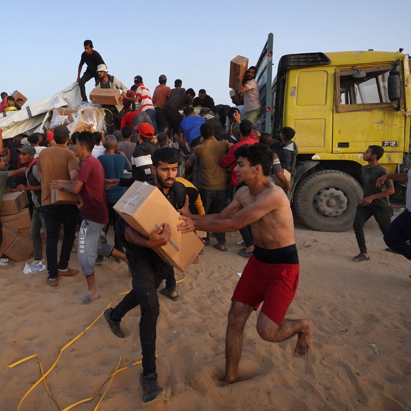 در این تصویر، گروهی از افراد در حال جابجایی و بارگیری جعبه‌ها از یک کامیون بزرگ هستند. برخی از افراد با جعبه‌ها به سمت کامیون می‌روند و دیگران در حال خارج کردن جعبه‌ها از کامیون به زمین هستند. به نظر می‌رسد این فعالیت در فضای باز و در یک منطقه شنی در حال انجام است. افراد در حال همکاری و تلاش برای حمل و نقل بسته‌ها هستند و جو تصویر به شدت忙 و پر از فعالیت به نظر می‌رسد.