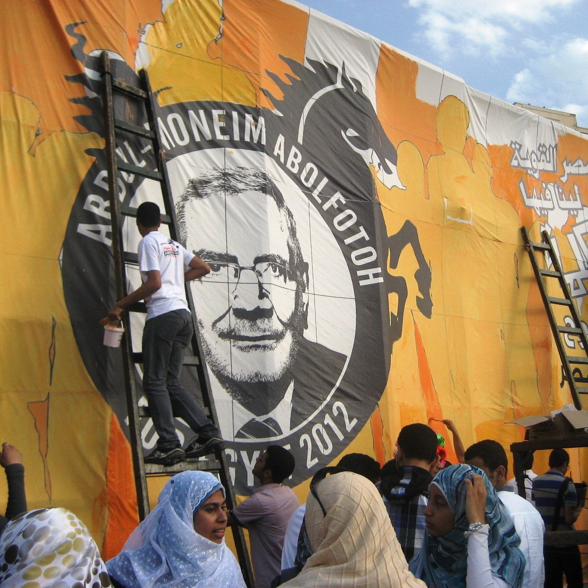 The image shows a group of people working together to paint or prepare a large mural or banner. The mural features a prominent circular design with the name "Abdel-Moneim Abol Fotoh" and the year "2012." There is a dragon motif in the background, which suggests a strong and bold theme. Some individuals are on ladders, actively engaged in applying paint, while others are gathered below, possibly helping or observing. The people are dressed in a variety of clothing styles, and there are some individuals wearing colorful wigs, indicating a festive or celebratory atmosphere. The backdrop is bright orange with various designs.
