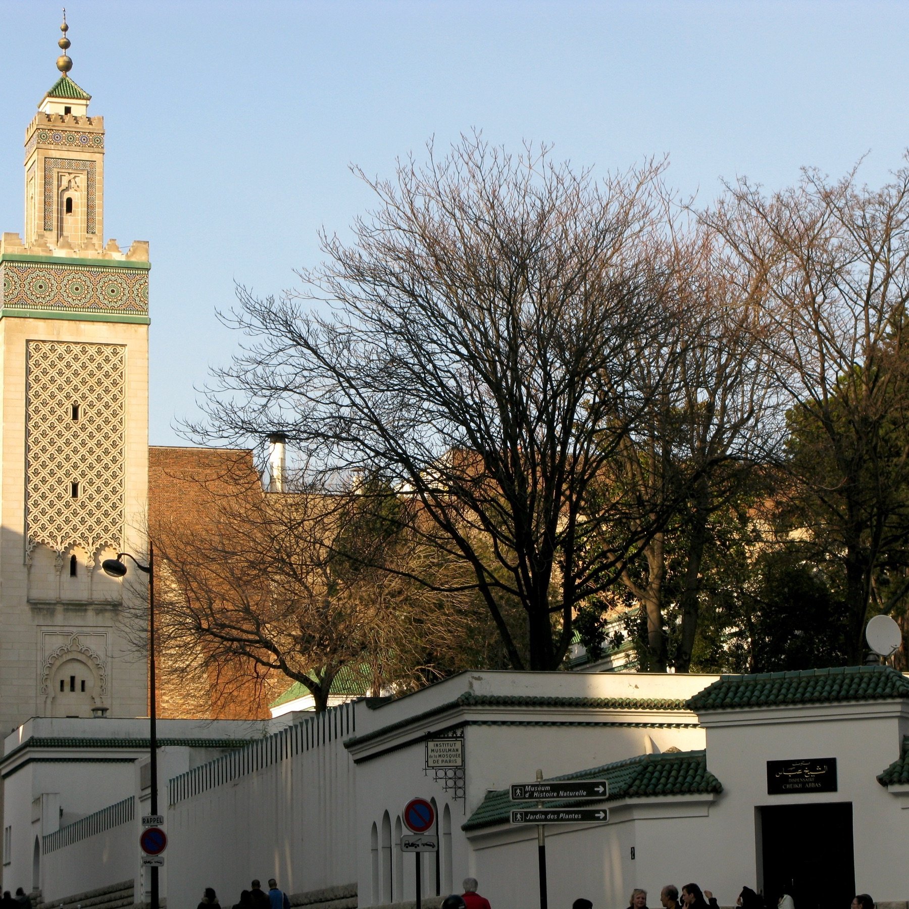 L'image montre une vue d'un bâtiment architectural distinctif, probablement une mosquée, avec un minaret élancé décoré de motifs. En arrière-plan, on aperçoit des arbres dépouillés, ce qui indique peut-être une saison hivernale ou automnale. Le mur entourant le bâtiment est blanc avec des bordures vertes. Des personnes marchent le long de la rue, créant une atmosphère de vie urbaine. Le ciel est dégagé, suggérant une journée ensoleillée.