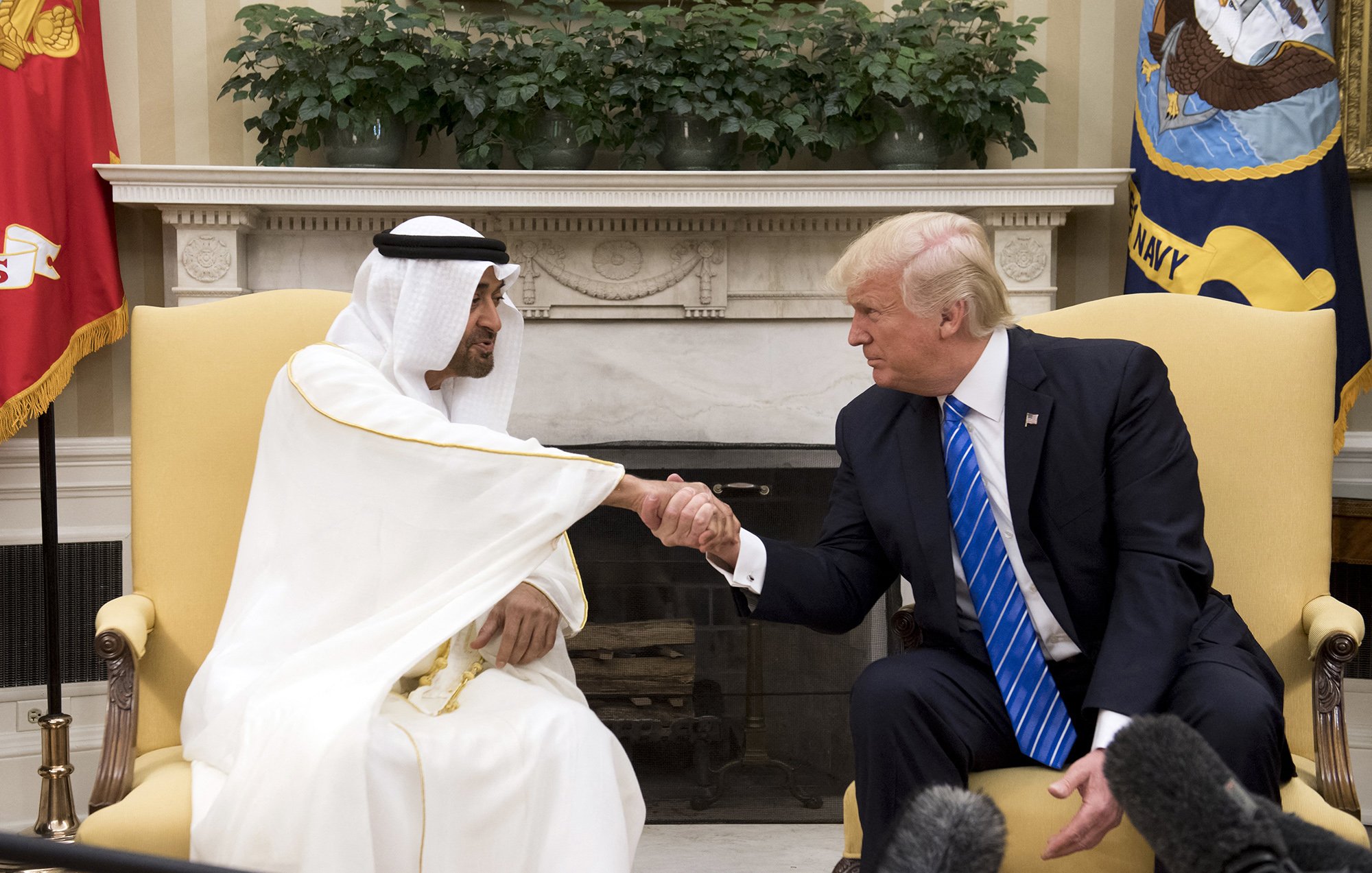The image shows a meeting between two men, one wearing a white traditional robe and headscarf, and the other in a suit with a blue tie. They are engaged in a handshake, indicating a friendly interaction. The setting appears to be an official office, possibly the White House, with flags and decorative elements in the background. There is a focus on their handshake, showcasing a moment of diplomacy.