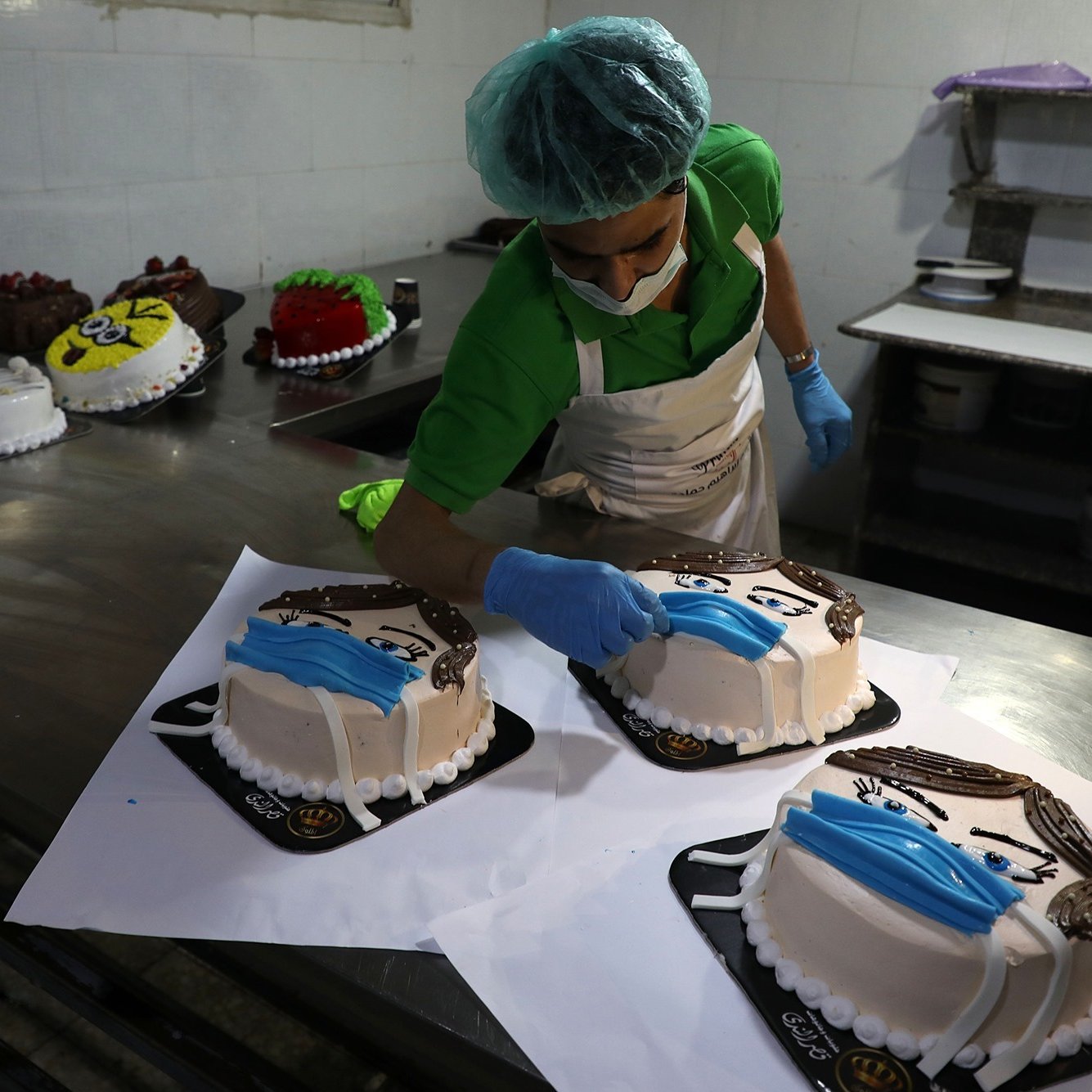 L'image montre un pâtissier en train de décorer des gâteaux dans une cuisine professionnelle. Il porte un bonnet de protection et des gants en latex. Sur la table, plusieurs gâteaux sont disposés, chacun ayant une décoration distincte, notamment des motifs qui ressemblent à des visages portant des masques. L'ambiance est soignée et les gâteaux sont présentés sur des plateaux noirs.