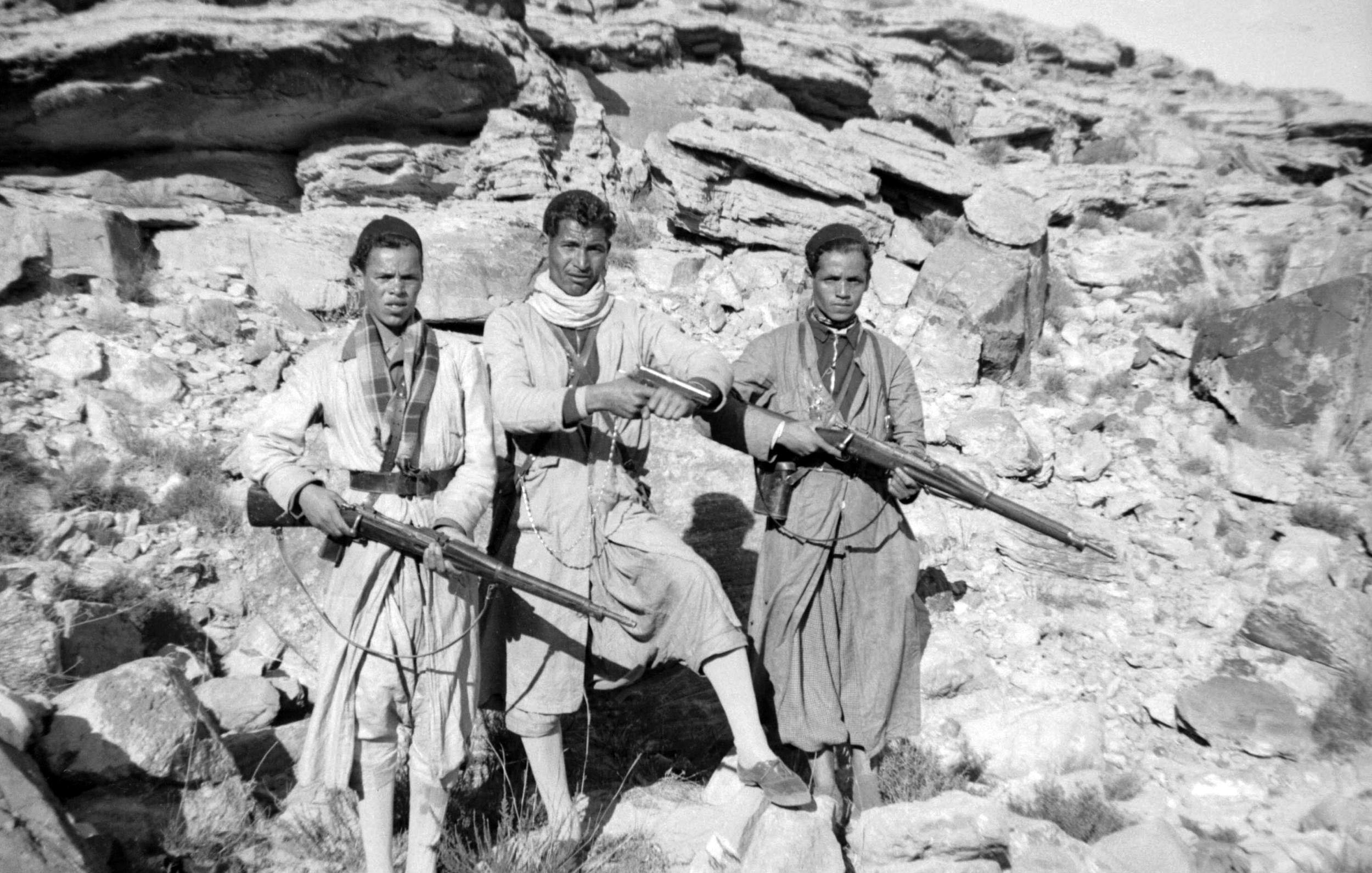 L'image montre trois hommes vêtus de vêtements traditionnels, tenant chacun un fusil. Ils se tiennent debout sur un sol rocheux, entourés de grandes pierres et de formations rocheuses. L'environnement semble austère et désertique. Les hommes ont des expressions sérieuses et sont alignés côte à côte. Leur posture et l'équipement qu'ils portent suggèrent qu'ils peuvent être des membres d'un groupe armé ou des guerriers appartenant à une communauté spécifique.