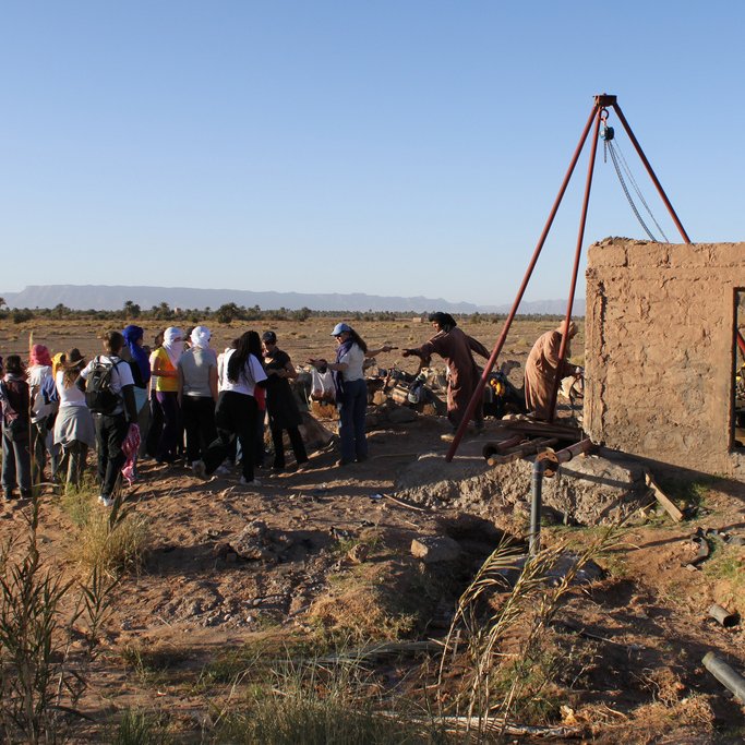 تظهر الصورة مجموعة من الأشخاص يتجمعون حول هيكل قديم في منطقة مفتوحة. الخلفية توضح منظرًا طبيعياً مع بعض التضاريس البعيدة مثل الجبال. الهيكل يبدو كأنه بناء غير مكتمل أو مهدم، ويمكن أن يكون جزءًا من مشروع أو نشاط معين. يبدو أن المشاركين يتابعون أو يتعلمون شيئًا ما متعلقًا بالهيكل. الأجواء واضحة ومشرقة، مما يضفي شعورًا بالحيوية على المشهد.