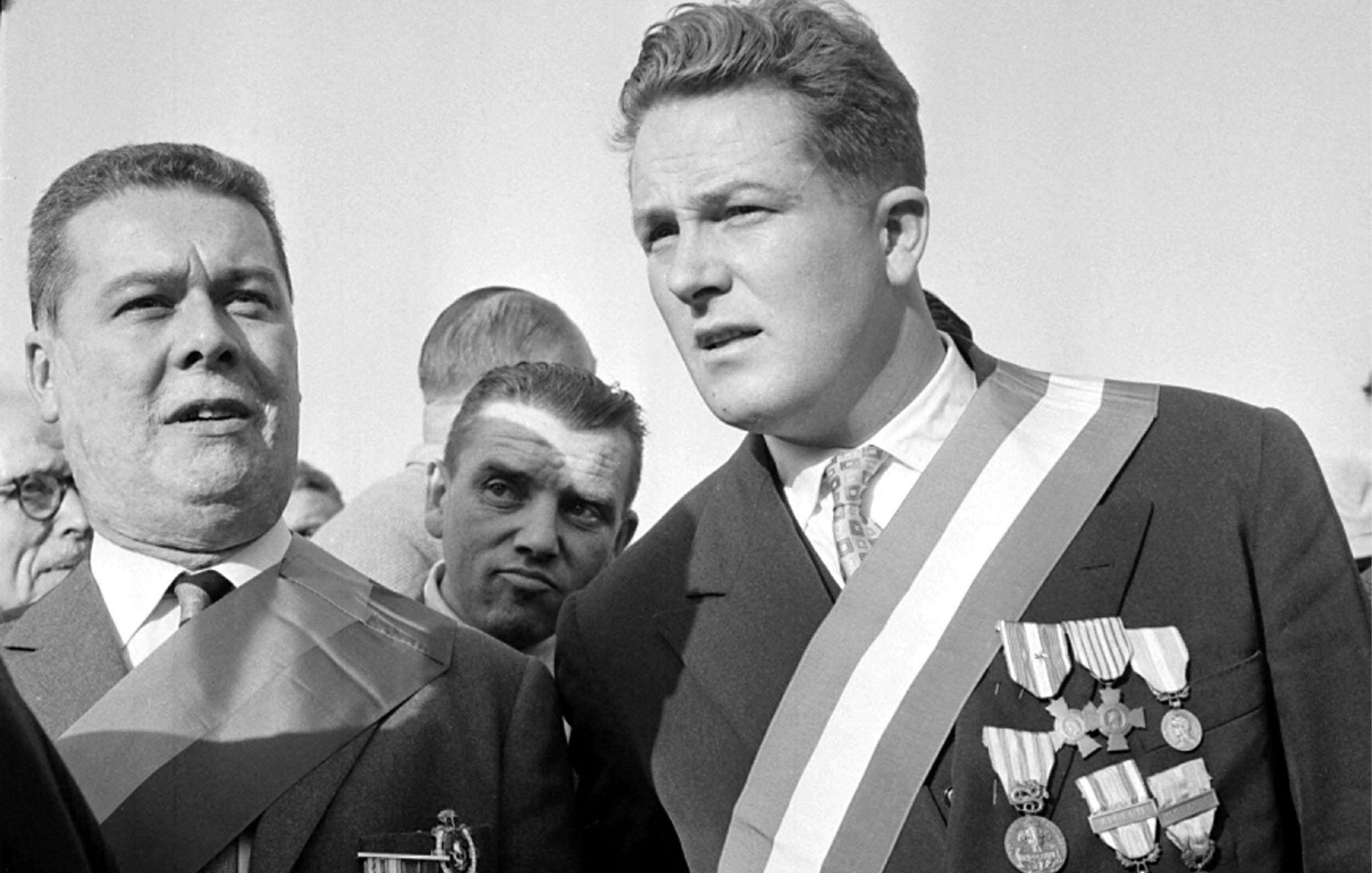 در این تصویر، دو مرد با لباس‌های رسمی و مدال‌های زیاد در حال صحبت یا مشاهده‌ی چیزی هستند. یکی از آن‌ها دارای مدال‌های متعددی بر روی سینه‌اش است و بر روی شانه‌اش نیز روبان بزرگی وجود دارد. مرد دیگر نیز به نظر می‌رسد مقام رسمی‌تری دارد و با دقت به طرف مقابل نگاه می‌کند. زمینه تصویر شامل افرادی دیگر است که به آنها توجه نمی‌شود. تصویر به وضوح فضایی رسمی و مربوط به یک مراسم یا رویداد مهم را نشان می‌دهد.