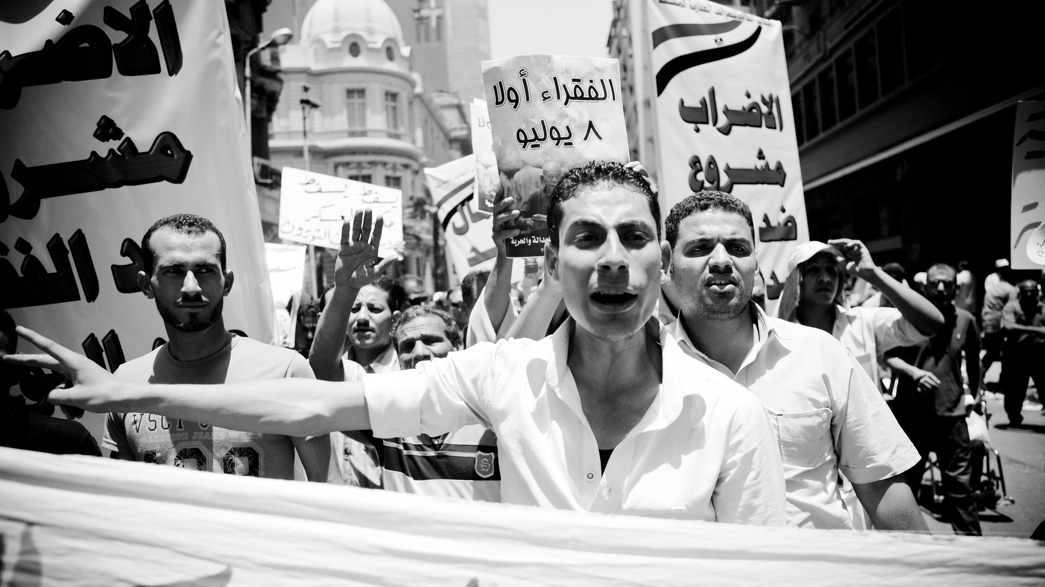 الصورة تُظهر مجموعة من المتظاهرين في شارع مزدحم، حيث يحملون لافتات ويهتفون بشعارات. يتقدم أحد المتظاهرين في المقدمة، ويبدو عليه الحماس. الجو يعكس أجواء التعبير عن المطالب والاحتجاج. تُظهر الصورة أيضاً لافتة كتب عليها "الإضراب الأول 8 يوليو"، مما يشير إلى موضوع الاحتجاج. الخلفية تحتوي على مباني حضرية، مما يضيف إلى السياق المدني للحدث.