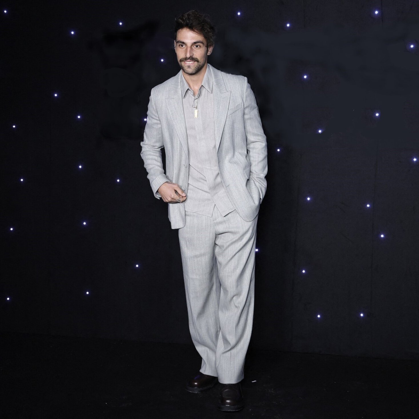 L'immagine mostra un uomo in piedi su uno sfondo scuro, illuminato da piccole luci bianche che brillano. Indossa un completo grigio chiaro, caratterizzato da pantaloni ampi e una giacca ben tagliata. La sua camicia è di un tono chiaro e ha il colletto aperto. L'uomo ha un sorriso e porta dei baffi, dando un'aria elegante e sicura di sé.