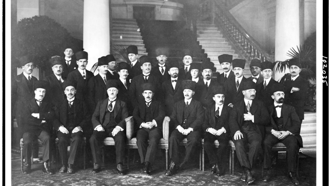 L'image présente un groupe d'hommes, probablement d'importance politique ou officielle, assis et debout dans un cadre formel. Ils portent des costumes sombres et des chapeaux traditionnels, et certains ont des moustaches. La scène se déroule dans un intérieur élégant, avec des colonnes décoratives et des plantes en pot en arrière-plan. L'atmosphère dégage une impression de solennité et de cérémonie, suggérant un événement ou une réunion importante.