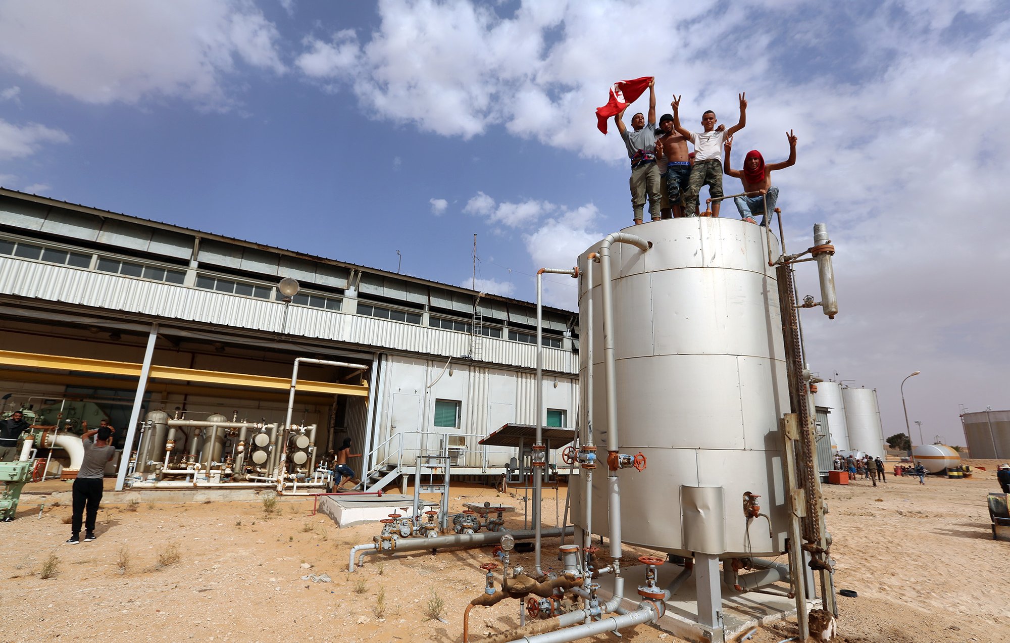 تظهر الصورة مجموعة من الأشخاص يقفون على قمة خزان معدني في منطقة صناعية، حيث يرفعون علمًا وينتشر خلفهم بعض المباني الصناعية. يبدو أنهم يحتفلون أو يعبرون عن فرحتهم، بينما يظهر في الخلف عدد قليل من الأشخاص الآخرين. السماء ملبدة بالغيوم، والأرض جافة. تعكس الصورة أجواء نشطة.