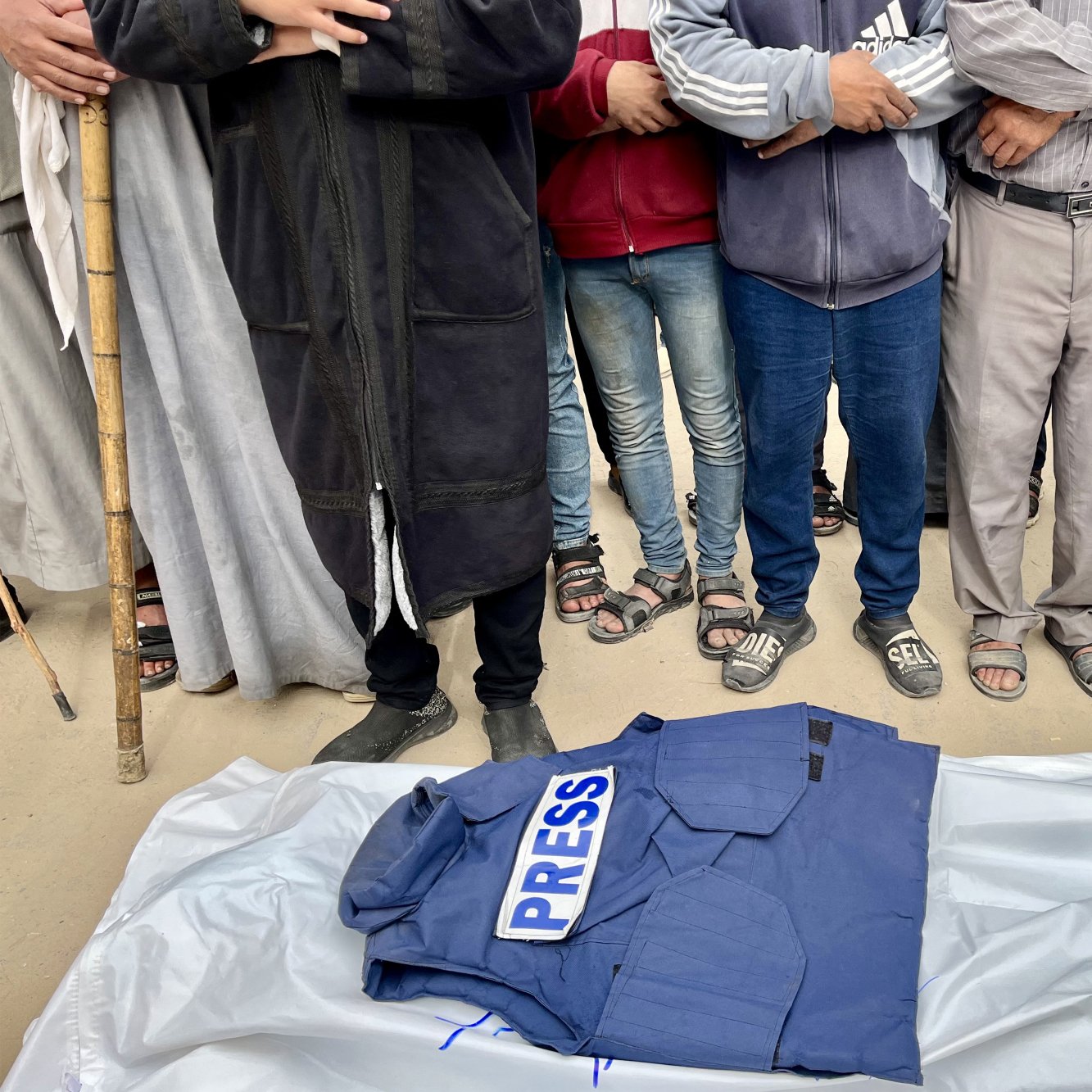A blue "PRESS" jacket rests on a white sheet, surrounded by people with arms crossed.