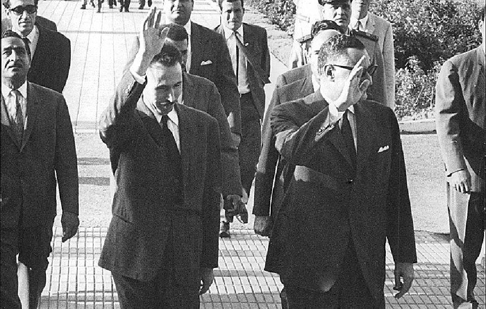 L'image montre un groupe de personnes marchant sur un chemin, avec plusieurs individus en costume. Deux hommes au premier plan saluent, vraisemblablement en se dirigeant vers une foule ou un événement. En arrière-plan, on peut apercevoir d'autres personnes et des éléments d'architecture, ainsi que des officiers en uniforme. L'atmosphère semble formelle et dynamique, indiquant une occasion spéciale.