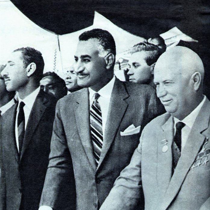L'image montre un groupe d'hommes en costume lors d'un événement officiel. Au centre, un homme avec une moustache et une cravate rayée est entouré de deux autres individus, dont l'un se tient à sa gauche. L'ambiance semble solennelle, et ils semblent être en train de discuter ou de participer à une cérémonie. L'arrière-plan montre une foule qui semble observer l'événement.