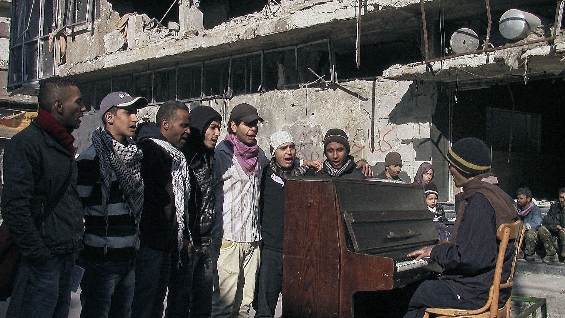 L'image montre un groupe de jeunes hommes chantant autour d'un piano dans un environnement dégradé, probablement après un conflit. Les bâtiments en arrière-plan sont en ruines, témoignant de destructions significatives. Les chanteurs semblent engagés et déterminés, tandis qu'un homme joue du piano, créant une atmosphère de solidarité et d'espoir au milieu de la désolation.
