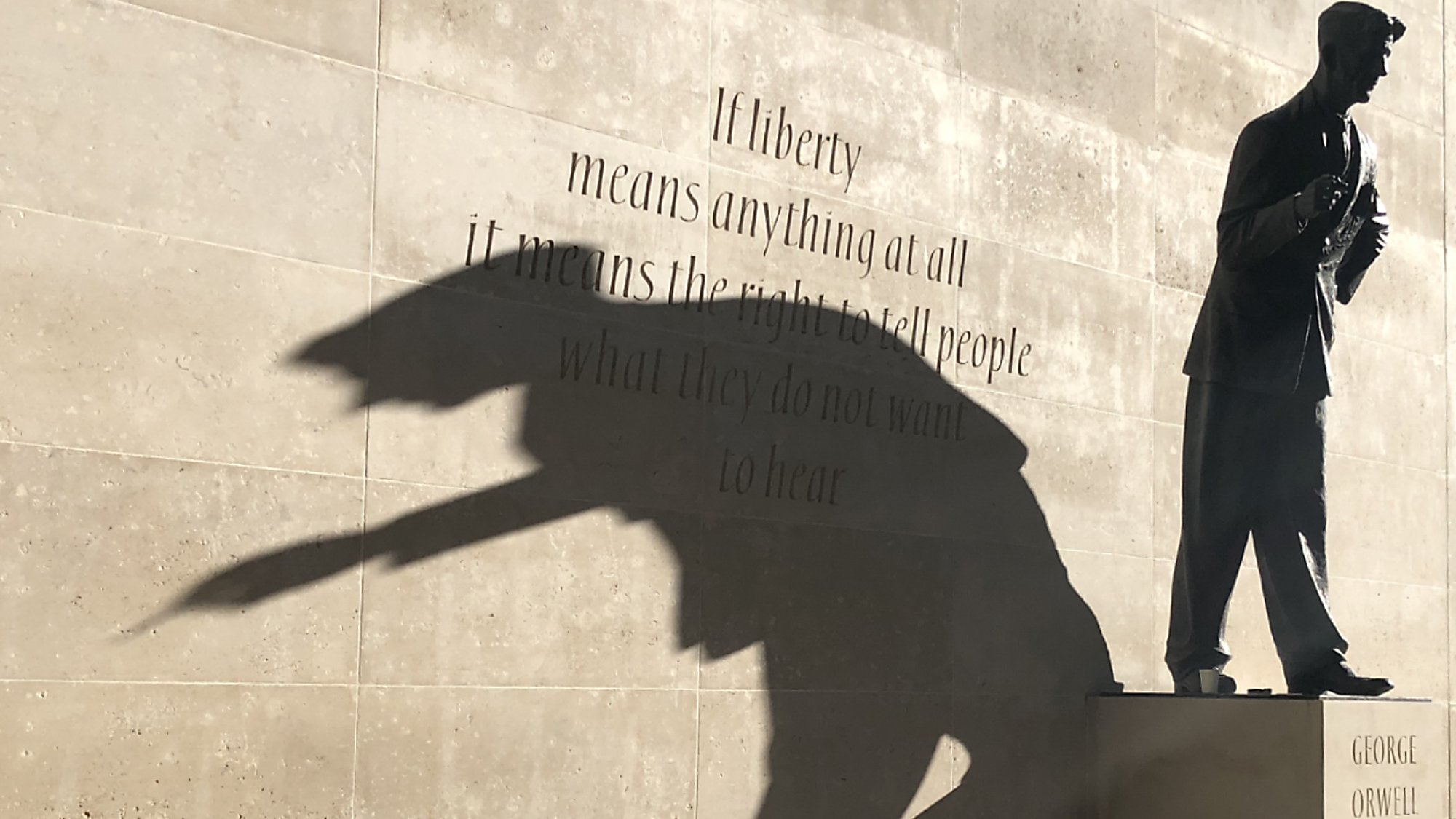 L'image montre une sculpture d'une silhouette d'un homme, probablement un écrivain, sur un socle avec le nom "GEORGE ORWELL". À côté de la statue, il y a une citation en anglais sur le thème de la liberté. La lumière crée une ombre qui semble projeter une forme dynamique, ajoutant une dimension artistique à la scène. Le mélange de texte et de sculpture évoque des réflexions sur la liberté d'expression.
