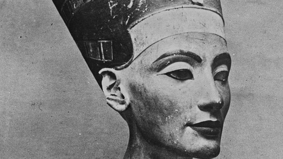 هذه صورة لتمثال نصفي لملكة أو إلهة مصرية قديمة، يُعتقد أنها نفرتيتي. يظهر التمثال النصفي بتفاصيل دقيقة وجميلة للوجه، بما في ذلك ملامح وجه متوازنة وعينين معبرتين. ترتدي الملكة تاجًا مميزًا مزينًا بتفاصيل دقيقة، بالإضافة إلى عقد رائع حول رقبتها. يُظهر التصميم دقة فنية تعكس الثقافة والفن المصري القديم.