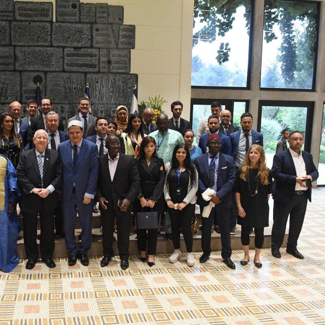 این تصویر گروهی از افراد را نشان می‌دهد که در یک مکان رسمی و با پس‌زمینه‌ای زیبا جمع شده‌اند. در وسط تصویر، چند مرد در کت و شلوار ایستاده‌اند که به نظر می‌رسد مسئولین یا مقام‌های بالاتر باشند. بقیه افراد، شامل مردان و زنانی با لباس‌های متنوع، در اطراف آنها ایستاده‌اند و همگی به دوربین نگاه می‌کنند. در پس‌زمینه، پرچم‌ها و یک دیوار زیبا مشاهده می‌شود که به محیط رسمی فضا افزوده است. افراد در این عکس به نظر می‌رسد که از ملیت‌ها و فرهنگ‌های مختلفی هستند.