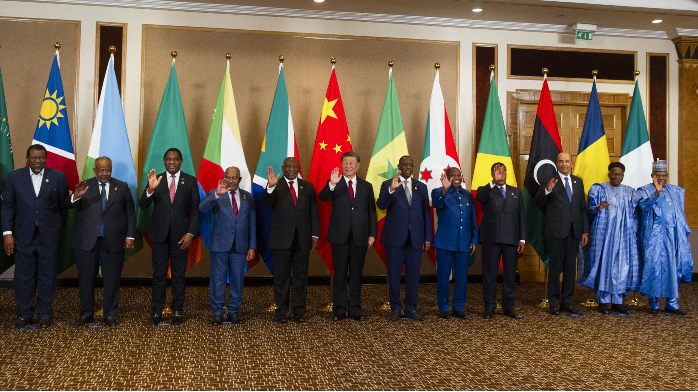 تصویر یک گروه از افراد را نشان می‌دهد که در کنار هم ایستاده‌اند و پرچم‌های مختلف کشورهایشان در پس‌زمینه قرار دارد. به نظر می‌رسد این افراد مقام‌های بلندپایه یا رهبران سیاسی هستند و همگی در حال خوش‌آمدگویی یا دست wave کردن به دوربین هستند. پرچم‌های مختلف شامل پرچم‌های کشورهای آفریقایی و همچنین پرچم‌های چین و سایر کشورها وجود دارد. فضای تصویر رسمی به نظر می‌رسد و افراد لباس‌های رسمی بر تن دارند.