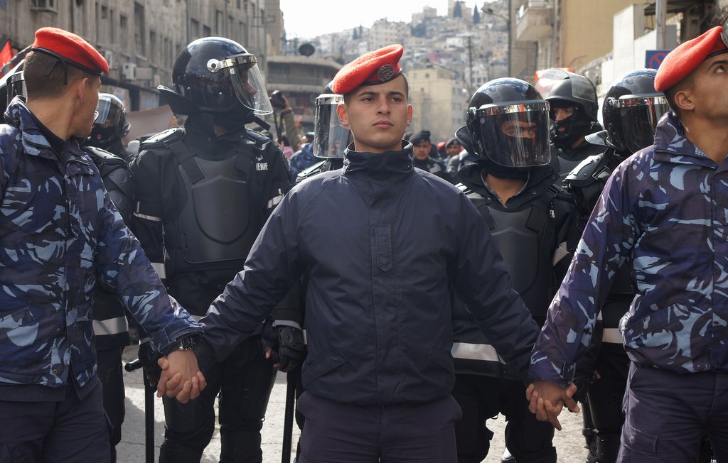 L'image montre un groupe de policiers ou de soldats, avec des uniformes distinctifs, se tenant les mains. Au centre, un homme en majorette avec un béret se distingue. Derrière eux, des membres des forces de l'ordre portent des casques et des équipements de protection. L'environnement semble urbain, avec des bâtiments en arrière-plan. L'atmosphère peut suggérer une situation de tension ou de manifestation.