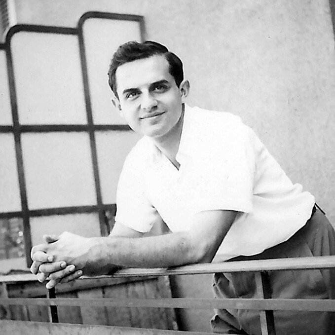 The image features a young man with a pleasant expression, leaning on a railing. He has dark hair styled in a neat manner and is dressed in a light-colored short-sleeve shirt paired with trousers. The background suggests a balcony or outdoor setting, with a partially visible modern-style partition or screen. The photo has a vintage quality, indicating it was likely taken several decades ago.