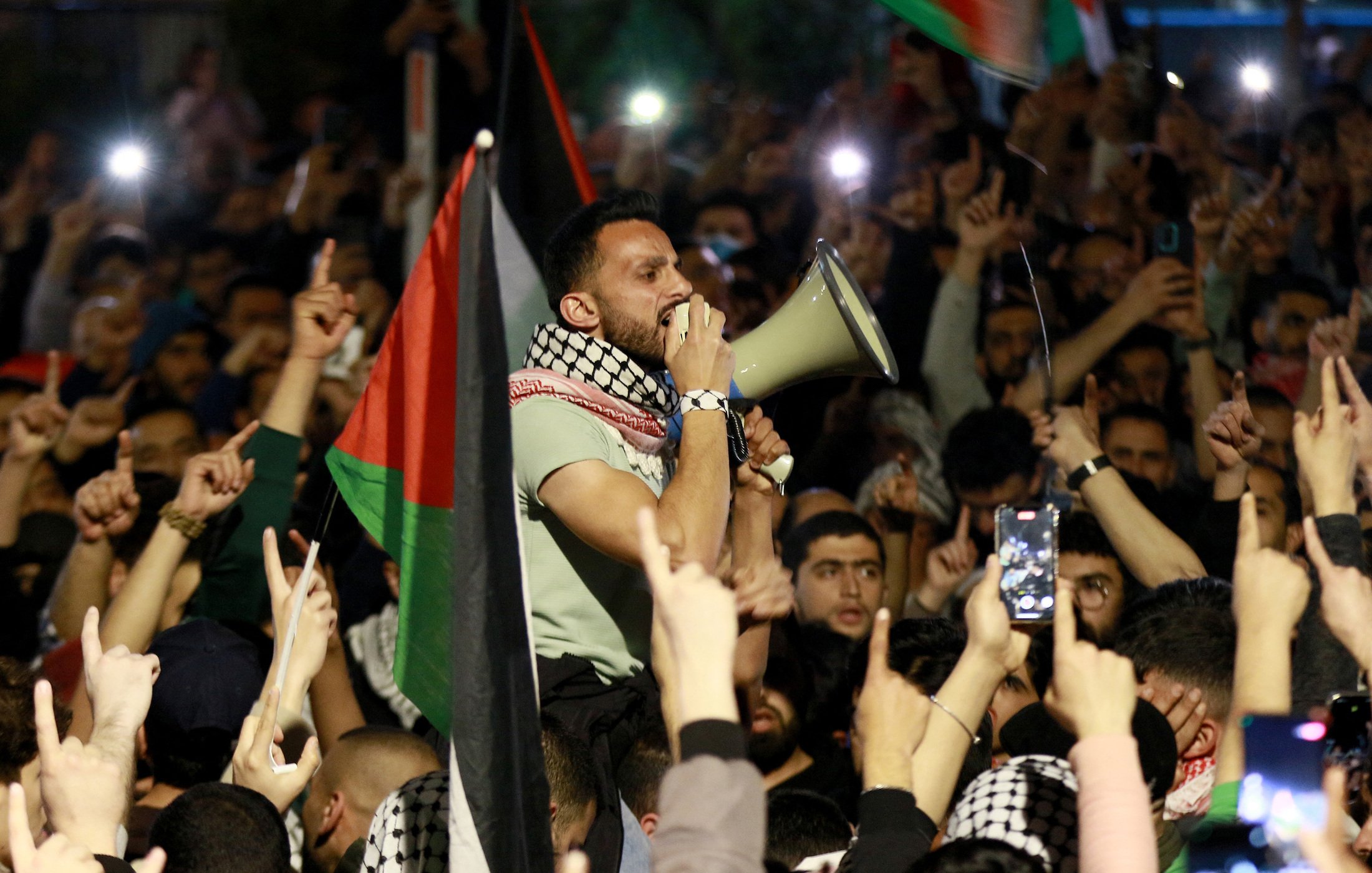 تصویر نشان‌دهنده جمعیتی از افراد است که در یک تجمع اعتراضی شرکت کرده‌اند. در مرکز تصویر، مردی با میکروفون در دست در حال سخنرانی یا فریاد زدن است. این فرد با پوششی که شامل شال معروف فلسطینی است، به نظر می‌رسد که به موضوعی مهم صحبت می‌کند. در پس‌زمینه، جمعیت زیادی با پرچم‌های فلسطین و دست‌هایی که به نشانه اعتراض یا حمایت بالا رفته‌اند، دیده می‌شود. این تصویر وضعیت هیجان‌انگیز و پرشور این تجمع را منتقل می‌کند.
