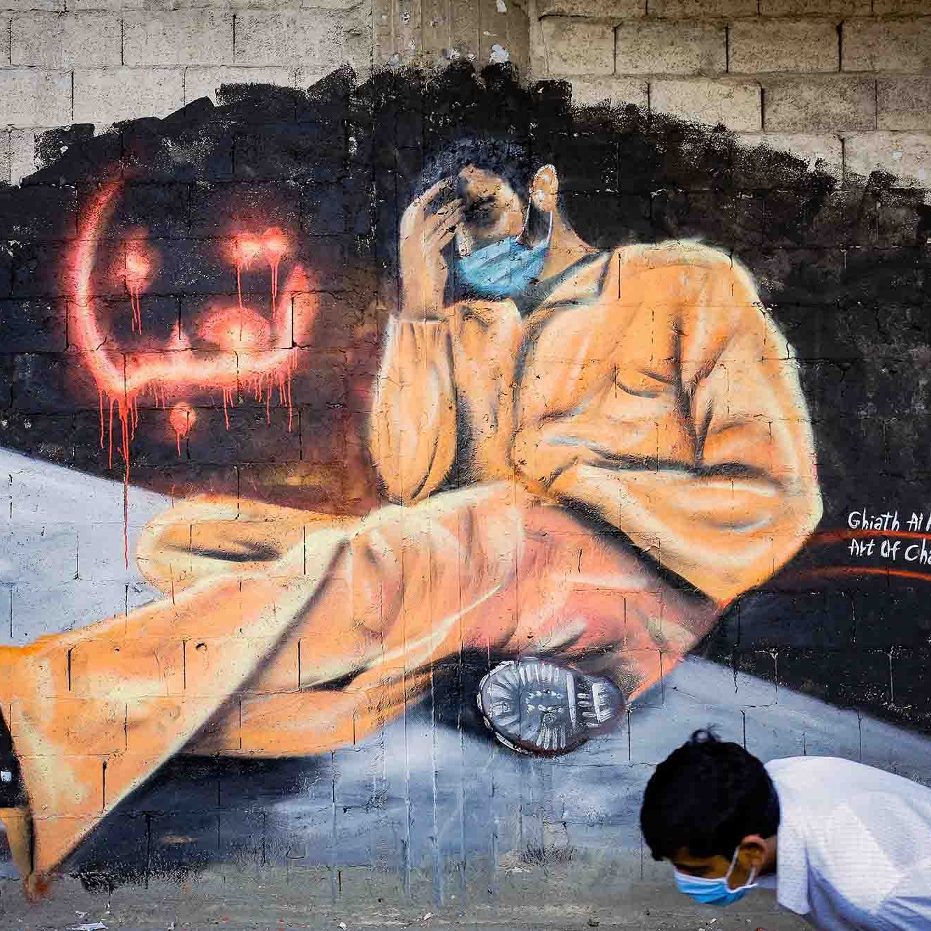 L'immagine mostra un murales che rappresenta una figura umana seduta, vestita con una tuta arancione, che tiene la testa tra le mani in un gesto di disperazione o riflessione. Il personaggio sembra essere in una posizione di vulnerabilità e solitudine. Accanto a lui, ci sono scritte in arabo e gocce di colore rosso che aggiungono un senso di drammaticità. Nella parte inferiore della scena, si vede un ragazzo in posizione inginocchiata, che sembra osservare il murales. Questo opera sembra voler trasmettere un messaggio profondo sulla sofferenza e l'oppressione.