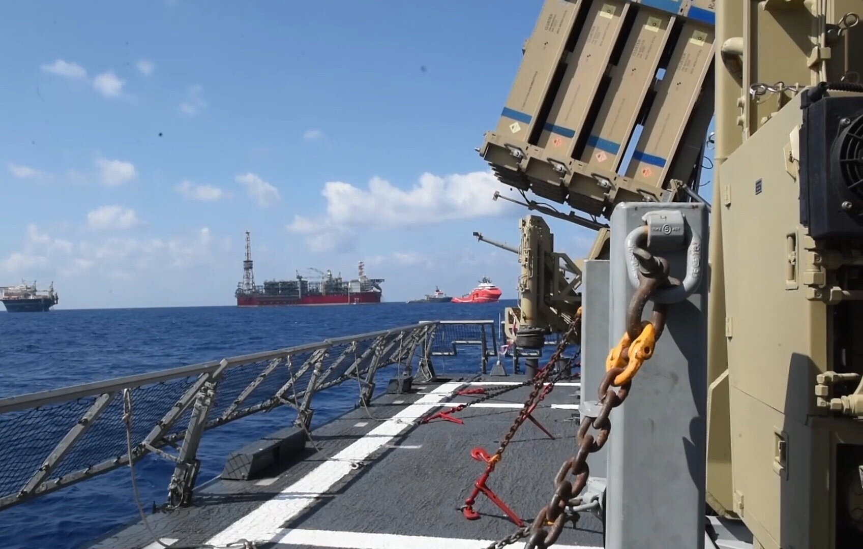 در تصویر یک نمای دریایی مشاهده می‌شود که شامل یک کشتی نظامی بخصوص با تسلیحات نصب شده بر روی آن است. در پس‌زمینه، چندین سکوی نفتی و کشتی دیگر در دریا دیده می‌شوند. آسمان آبی و روشن است و چند ابر نیز در آن وجود دارد. فضای کلی تصویر به نظر می‌رسد که در حال اجرای عملیات دریایی یا امنیتی است.