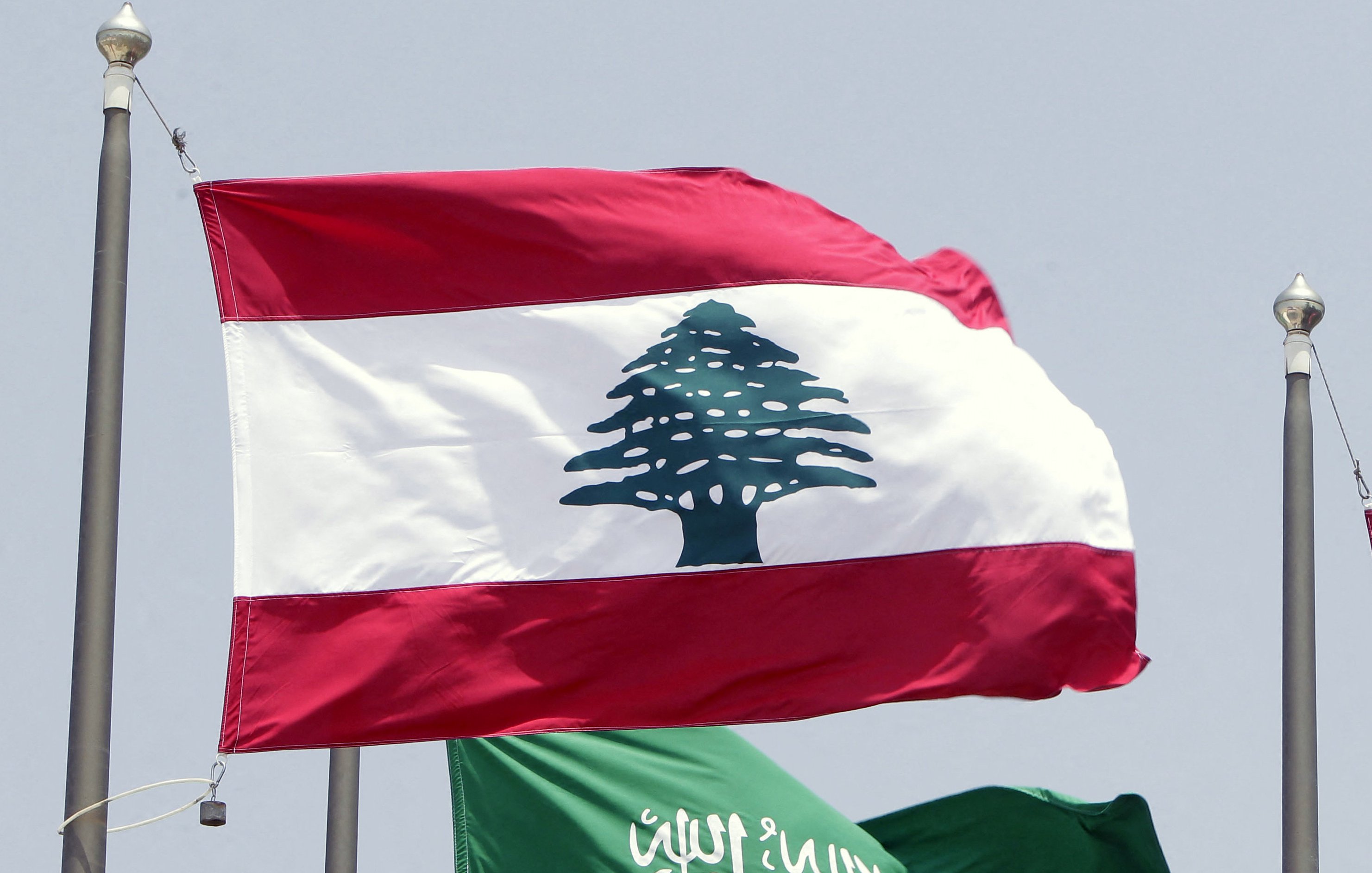 L'image montre deux drapeaux flottant côte à côte. Le drapeau du Liban est en haut, avec une bande rouge en haut et en bas, et une grande illustration d'un cèdre vert au centre. En dessous, le drapeau de l'Arabie Saoudite est vert, portant une inscription en arabe et une épée blanche. Les drapeaux sont hissés sur des mats, probablement lors d'un événement officiel ou d'une rencontre diplomatique.