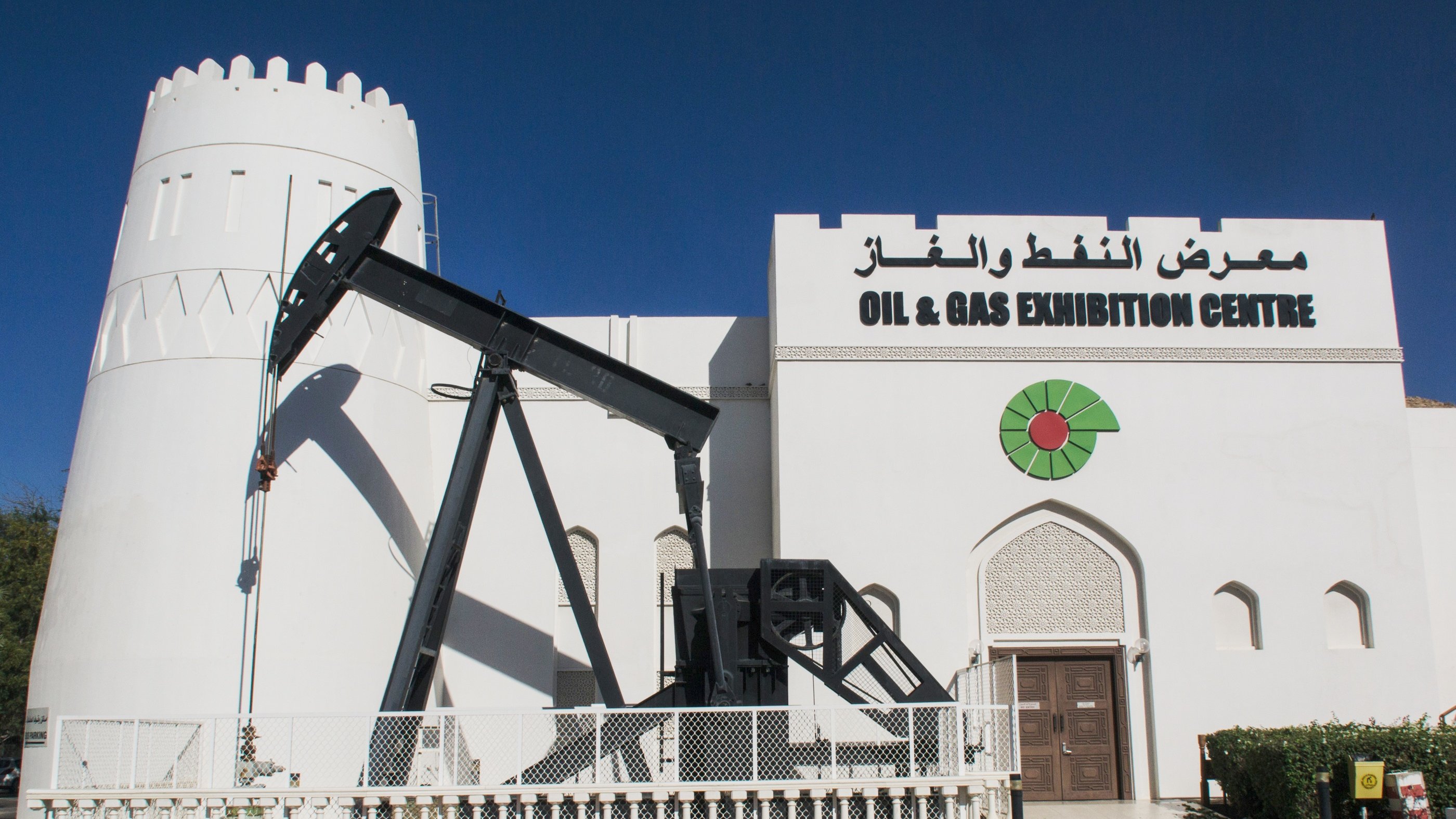 The image features a white, castle-like building with a prominent sign that reads "OIL & GAS EXHIBITION CENTRE" in both English and Arabic. To the left of the entrance, there is an oil pump jack, a piece of machinery commonly used in oil extraction. The building has a decorative design with a combination of traditional and modern architectural elements. The sky is clear and blue, creating a vibrant backdrop.