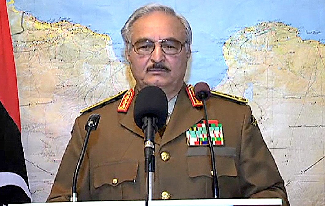 L'image montre un homme portant un uniforme militaire, avec plusieurs médailles et distinctions sur sa veste. Il se tient derrière un pupitre de conférence, devant une carte géographique en arrière-plan. L'homme semble s'adresser à un public ou à une caméra. On peut voir des drapeaux en arrière-plan, indiquant une situation officielle.