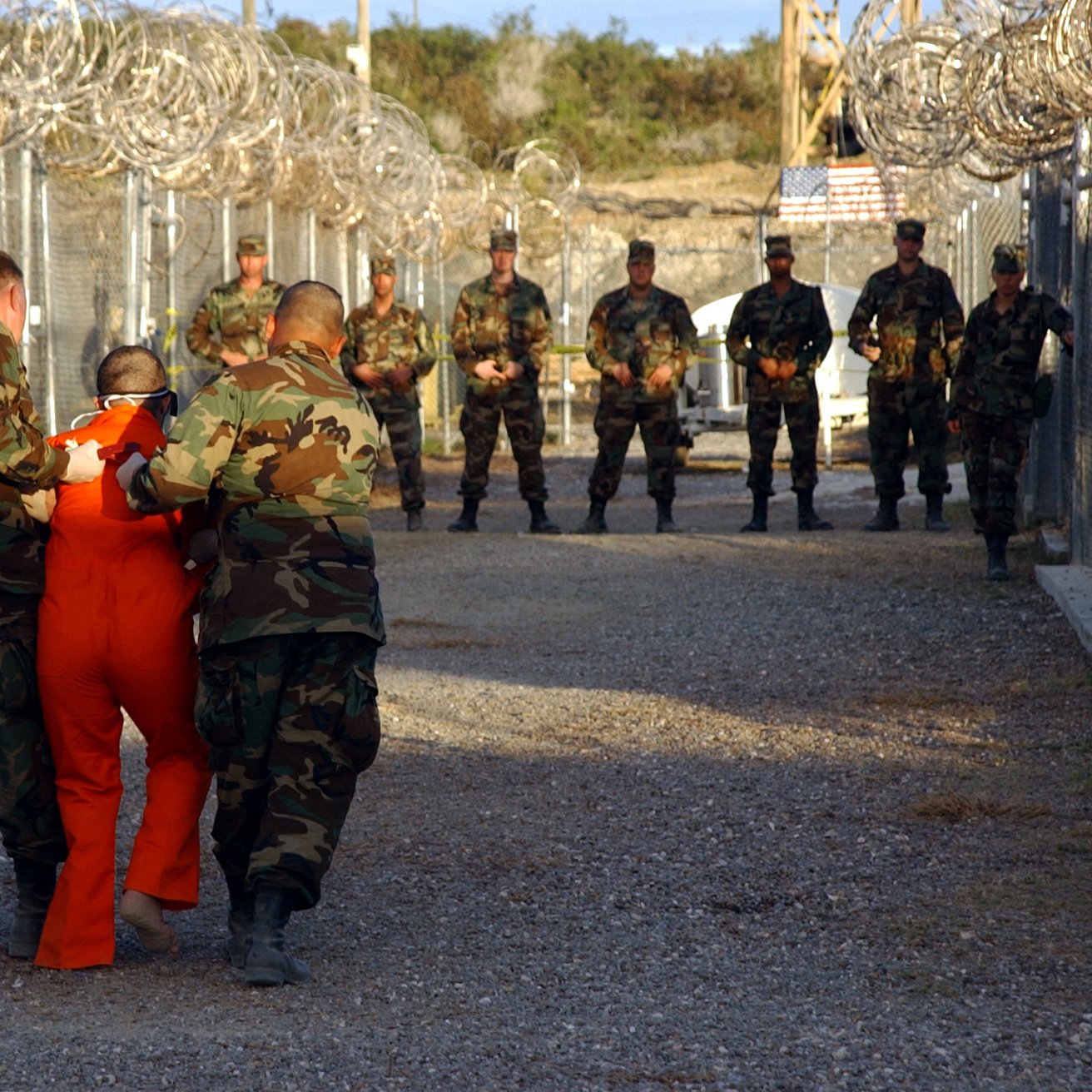 در این تصویر، چند نظامی در لباس‌های نظامی در یک فضای محصور با دیوارهای مشبک و نوارهای خاردار حضور دارند. یکی از نظامیان در حال هدایت یک فرد در لباس نارنجی است که به نظر می‌رسد دستگیر شده باشد. تعدادی دیگر از نظامیان در پس‌زمینه ایستاده‌اند و به صحنه نظاره می‌کنند. فضا به شکل یک محوطه امنیتی یا زندان به نظر می‌رسد.