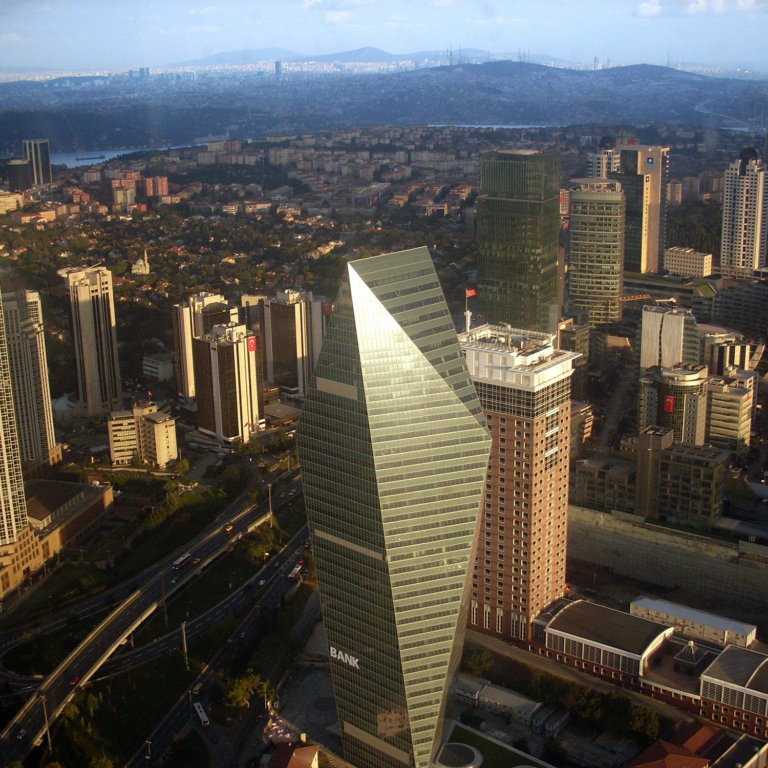 The image depicts a cityscape with a collection of modern skyscrapers and buildings. In the foreground, a distinctive triangular-shaped building stands out, likely serving as a bank or a financial institution. Surrounding it are various other high-rise buildings, indicating a bustling urban environment. The scene appears to be taken from a high vantage point, as it provides a wide view of the city, including green areas and roads. The skyline is clear, suggesting a bright day, with distant mountains or hills visible in the background.