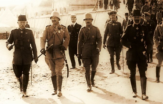 The image depicts a historical scene with a group of military personnel walking together in a line. They are dressed in formal military uniforms and accessories, including hats and medals, indicating a high rank or importance. The setting appears to be outdoors, possibly on a dirt or gravel path, with tents in the background suggesting a military camp. The overall tone of the photograph has a sepia or monochrome effect, conveying a sense of history. The individuals appear to be engaged in conversation or in a social setting, reflecting camaraderie among them.