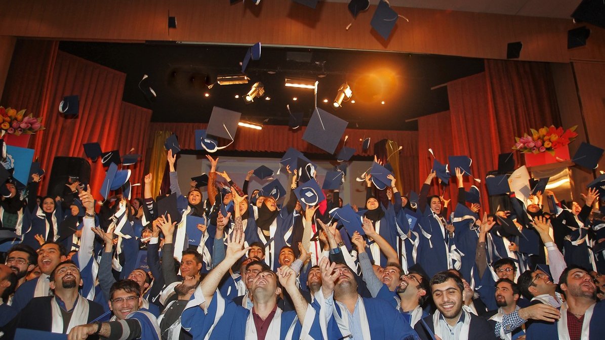 L'image montre une célébration de remise de diplômes. De nombreux étudiants, vêtus de toges et de mortiers de graduation, se tiennent ensemble sur scène. Ils lèvent leurs chapeaux en l'air, exprimant leur joie et leur excitation. L'atmosphère est festive, avec des sourires sur les visages des diplômés, soulignant un moment marquant de leur parcours académique. Des lumières et des décorations de scène ajoutent à l'ambiance festive.