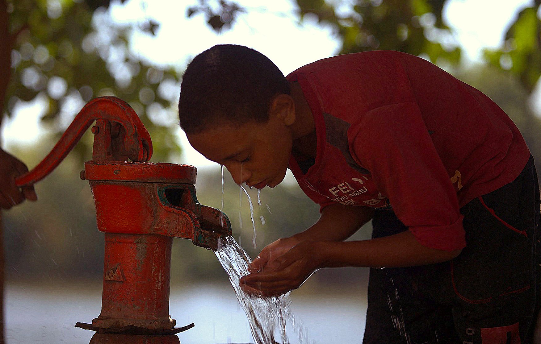 The image shows a young boy bent over a red hand pump, drawing water from it. He appears focused as he cups his hands to catch the water flowing out. The background features greenery and suggests a natural setting, possibly near a body of water, with soft light creating a warm atmosphere. The boy is wearing a red shirt, and the scene conveys a sense of simplicity and the importance of access to fresh water.